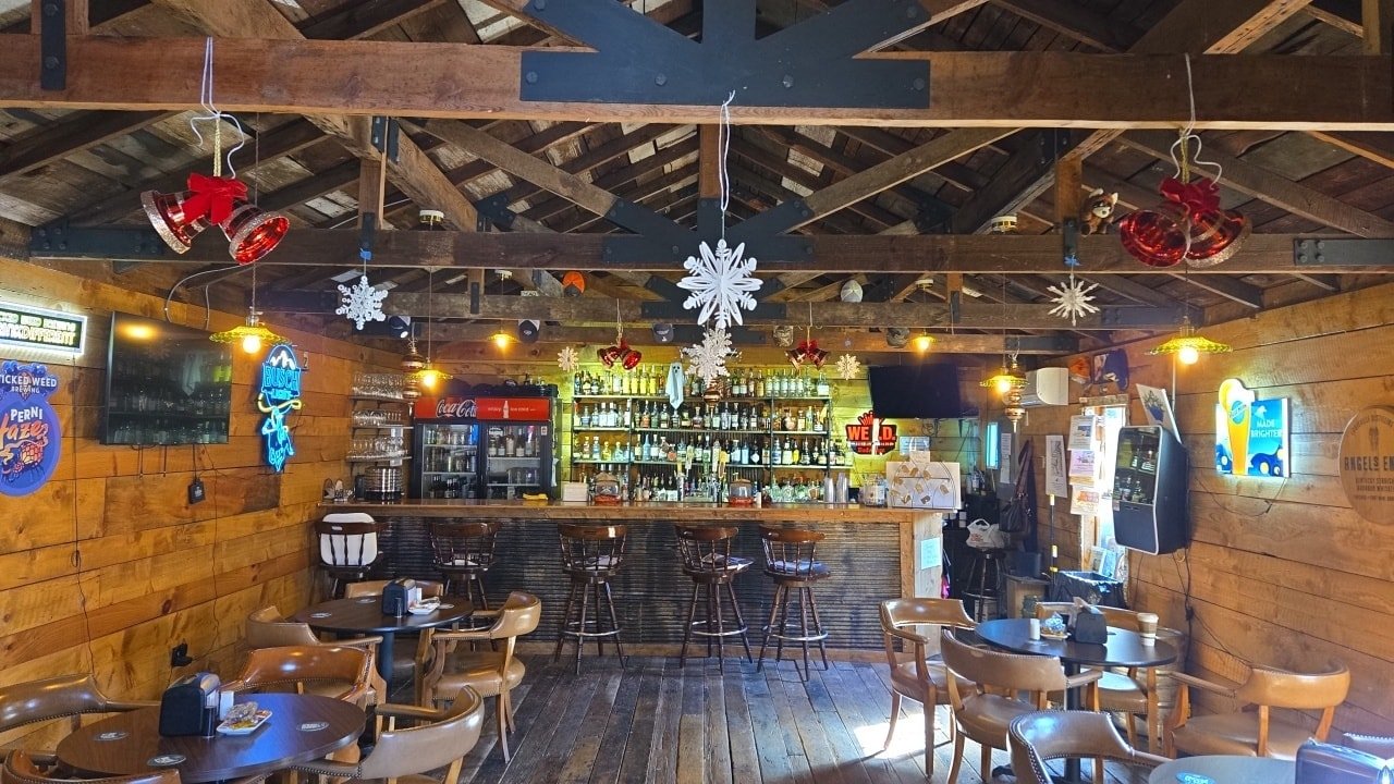 Inside a rustic bar with wooden walls and ceiling, decorated with hanging snowflakes and red jingle bells, featuring a bar counter with bar stools, a TV, neon signs, and various bottles of alcohol on shelves behind the bar.