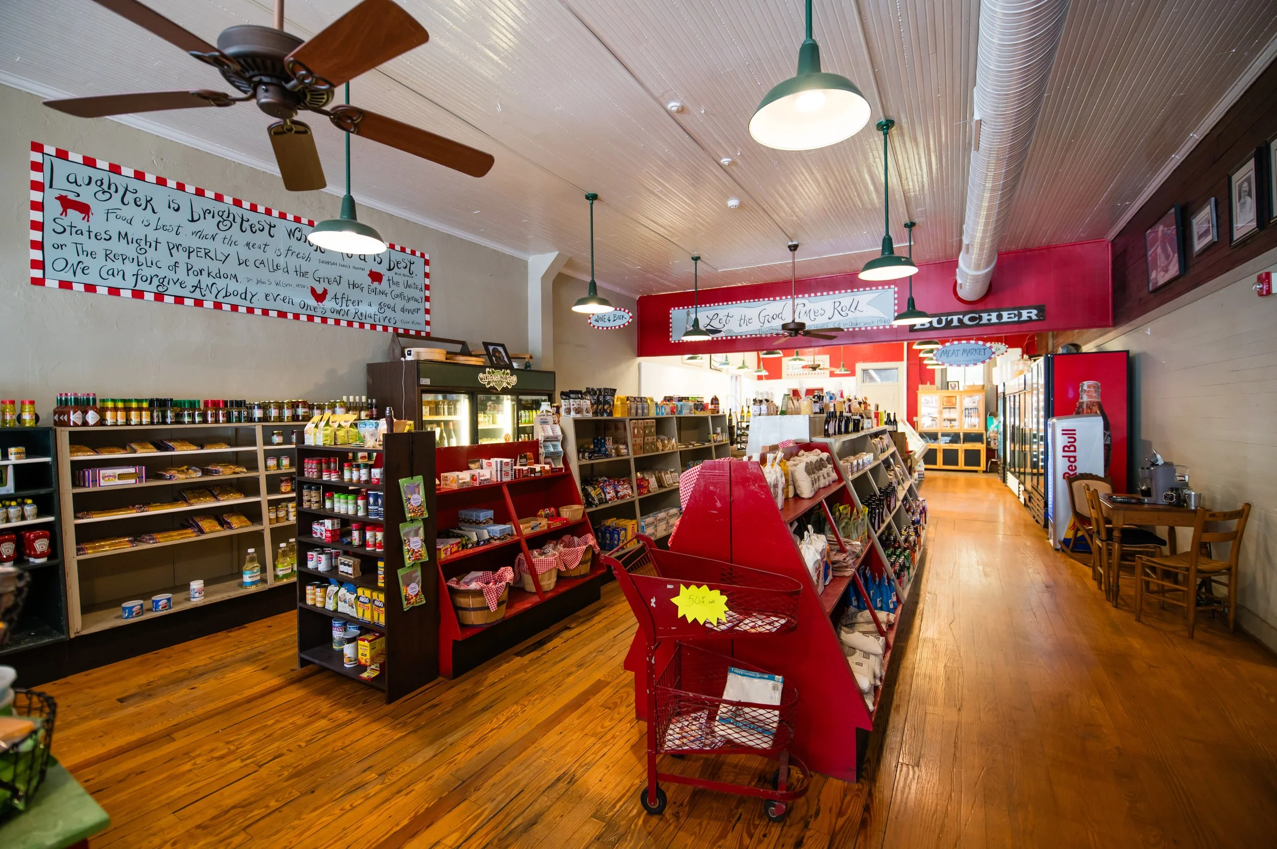 Interior of a grocery or general store with wooden floors, shelves filled with canned goods, snacks, and other products, a red shopping cart, ceiling fans, hanging lights, and framed photos on the wall.