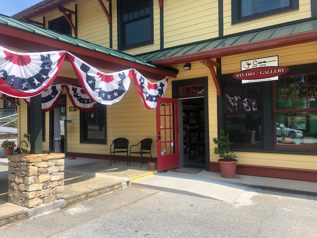 Exterior of a gallery or studio with red door, black chairs, potted plants, patriotic bunting, and a sign that reads 'Studio / Gallery'.