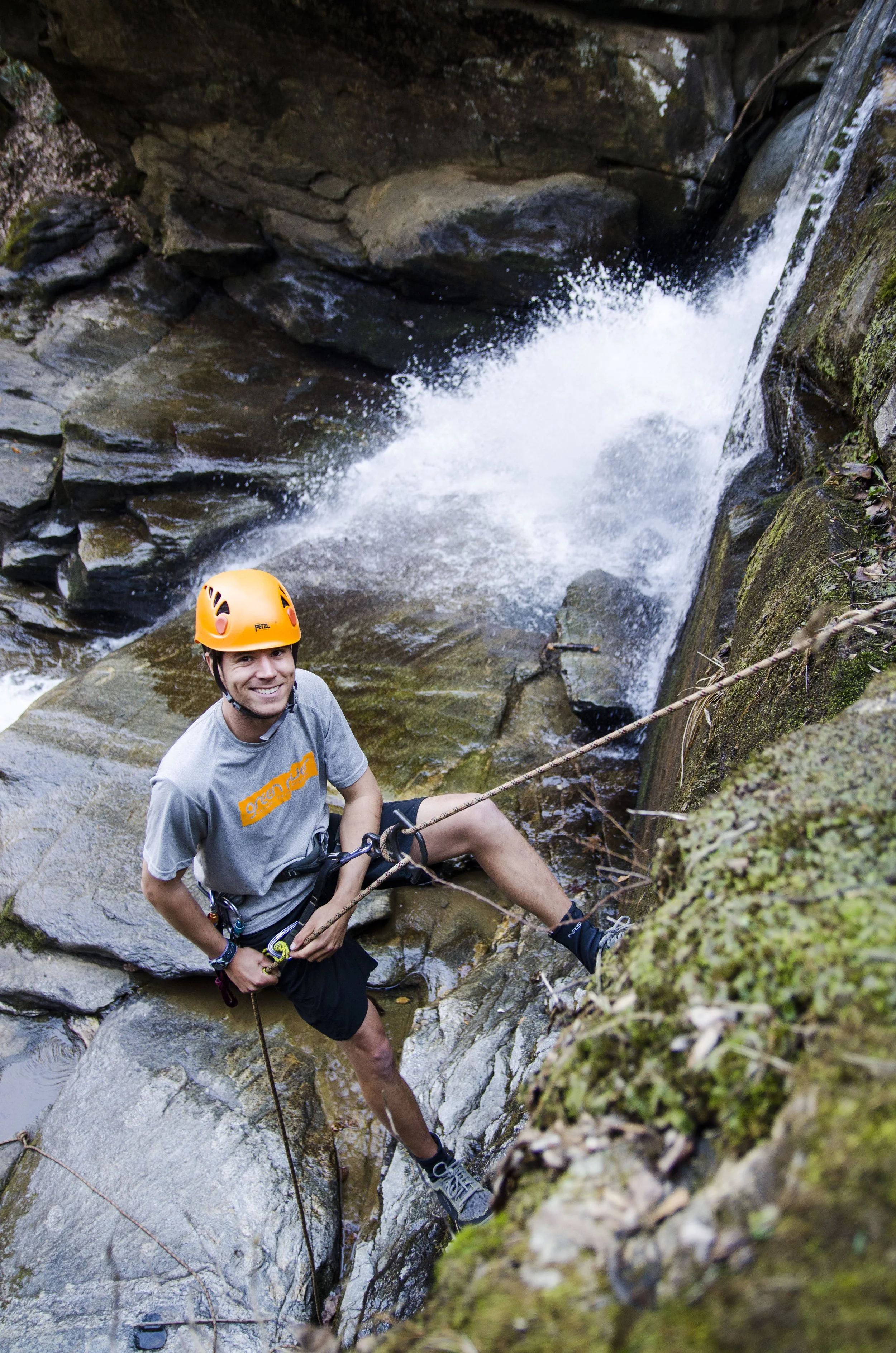 A man in climbing gear, wearing a yellow helmet and grey T-shirt, standing on a rocky surface next to a waterfall or rapid stream in a forested area.