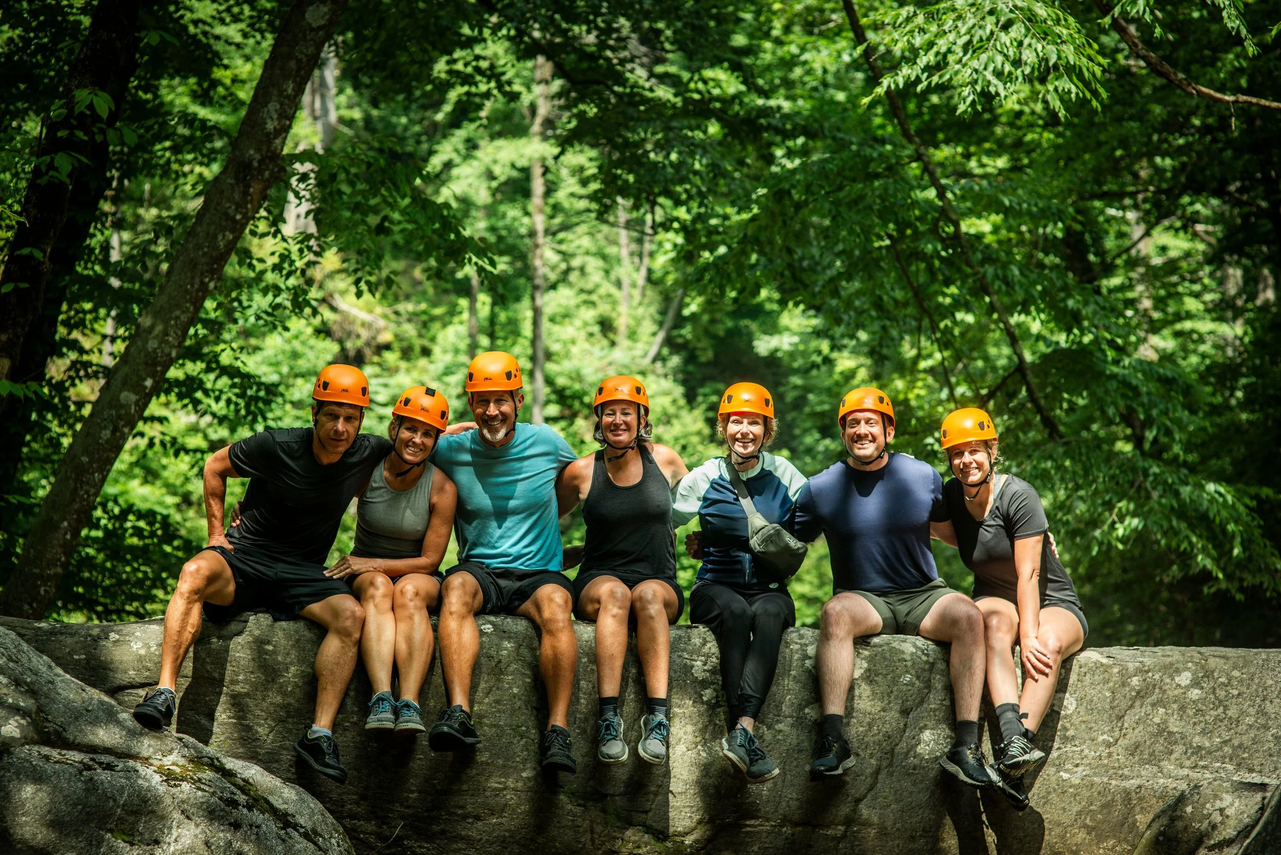 A group of seven people wearing safety helmets sitting on a tree log in a forest.