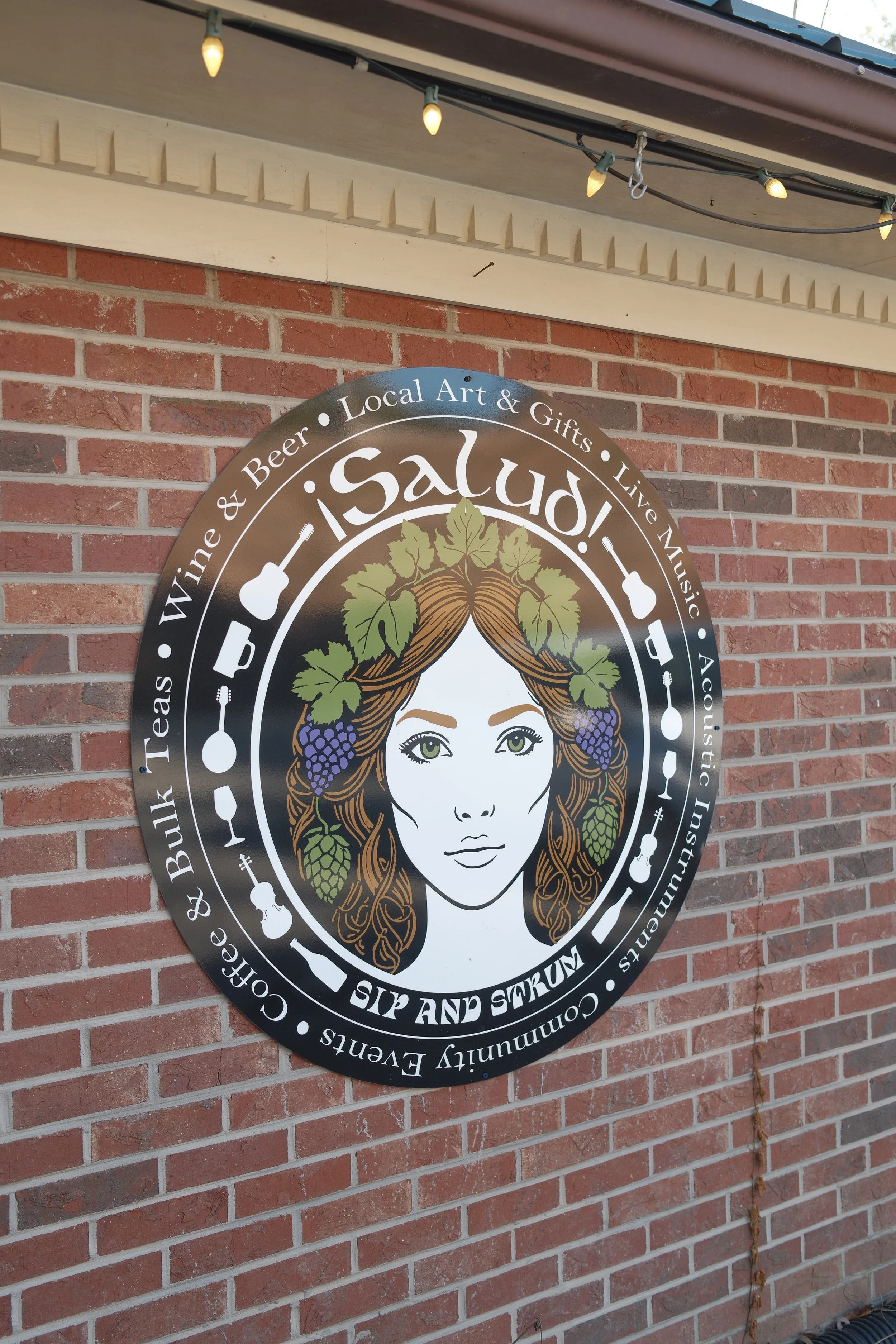 A circular sign on a brick wall promoting a business called 'Salud' that sells wine, beer, local art, gifts, and live music. The sign features an illustration of a woman with hair adorned with grapevine leaves and grapes, with decorative icons of mus