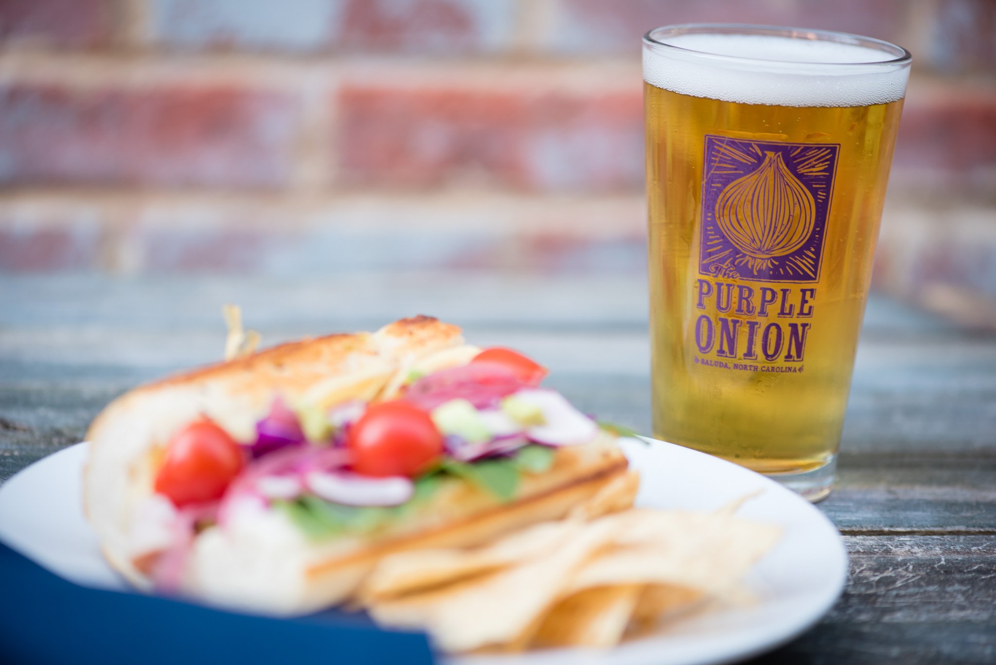 A glass of beer with a purple and yellow logo on it that says 'The Purple Onion' and a plate of sandwich with vegetables in the foreground, all on a rustic wooden table with a blurred brick wall background.