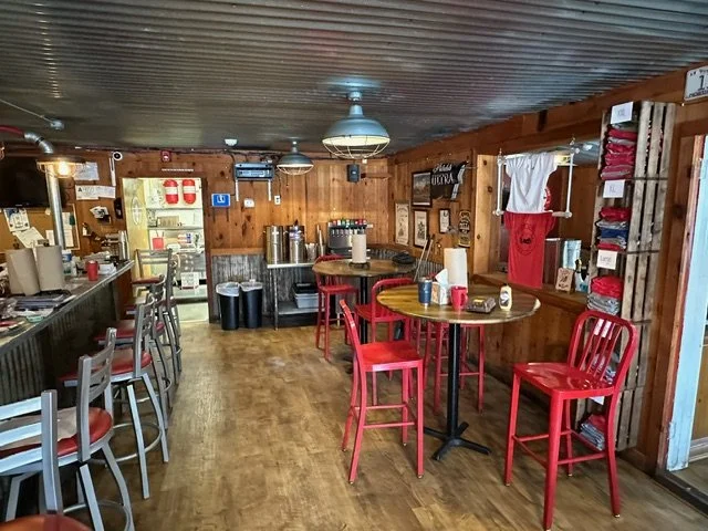 Interior of a rustic restaurant or cafe with wooden walls, red chairs, and a bar area on the left side with bar stools. There are tables with condiments and napkins, and various items hanging on the walls, including T-shirts and signs.
