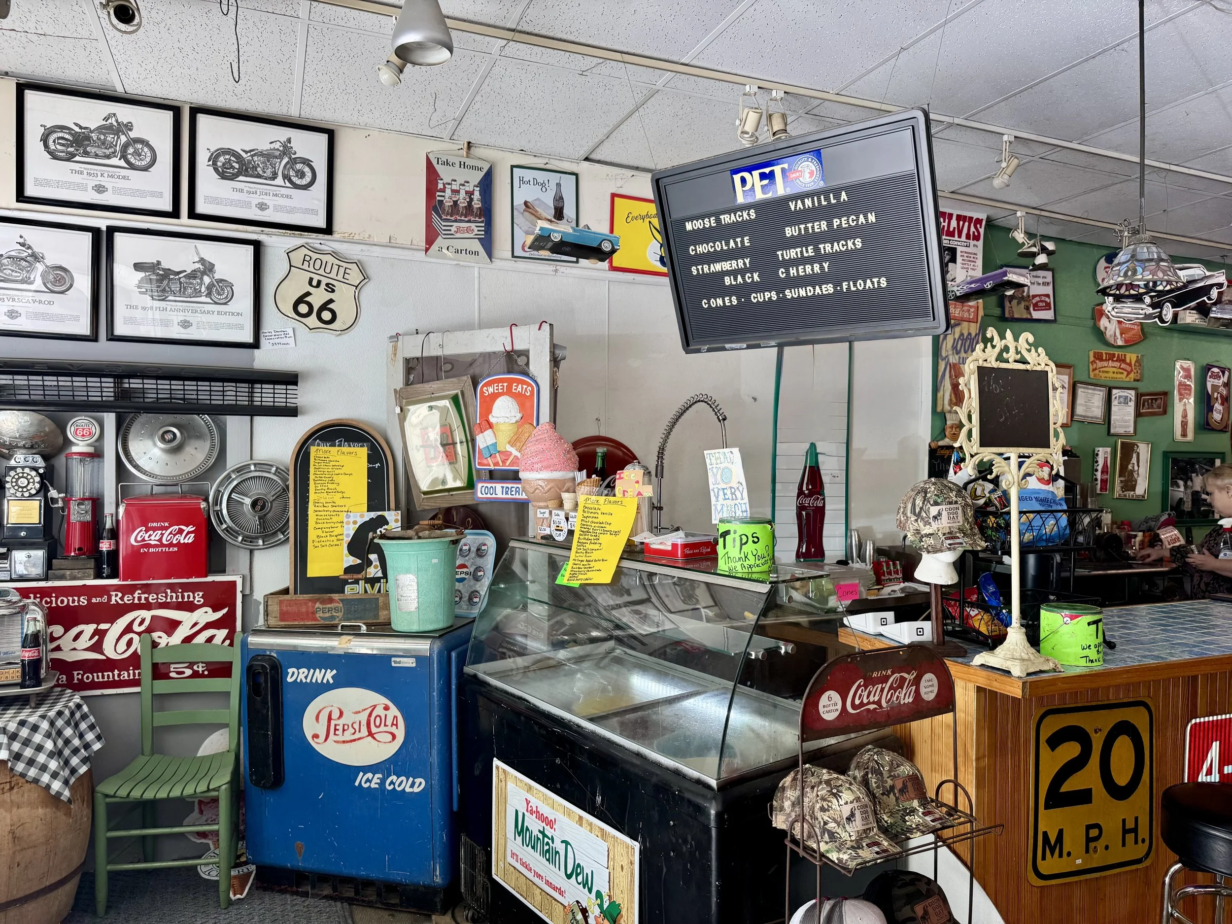 Interior of a vintage diner or cafe with framed motorcycle pictures, a Route 66 sign, a menu board listing various pet ice cream flavors, Coca-Cola decorations, and retro decor items.