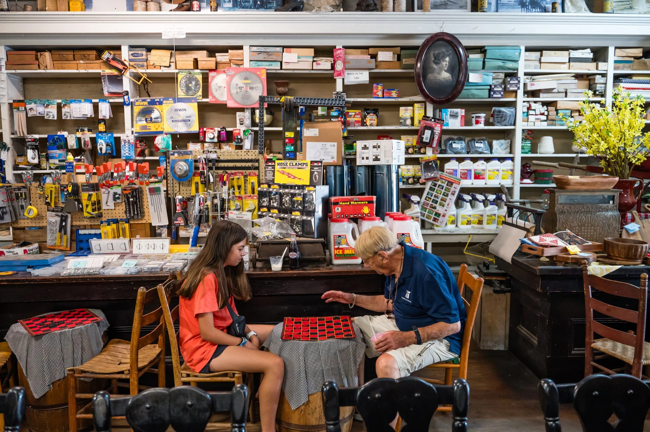 A vintage-style store with shelves of supplies and organized items, two people playing checkers at a table, and two children with an elderly woman in the foreground.