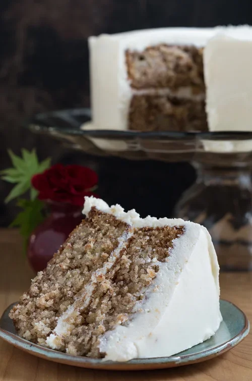 Slice of layered cake with white frosting on a small plate in the foreground, with a cake stand holding more slices of the same cake in the background.