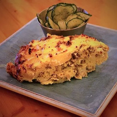 A slice of casserole with a baked topping served on a blue plate, accompanied by a bowl of sliced pickles.