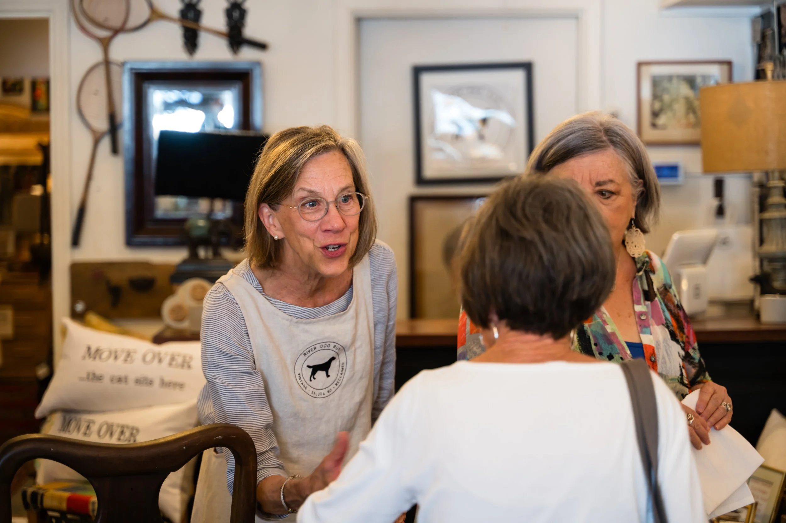 Three women engaging in conversation around a table in a cozy, decorated room with framed artwork and hunting trophies on the wall. A pillow with the text 'Move Over' is visible in the foreground.