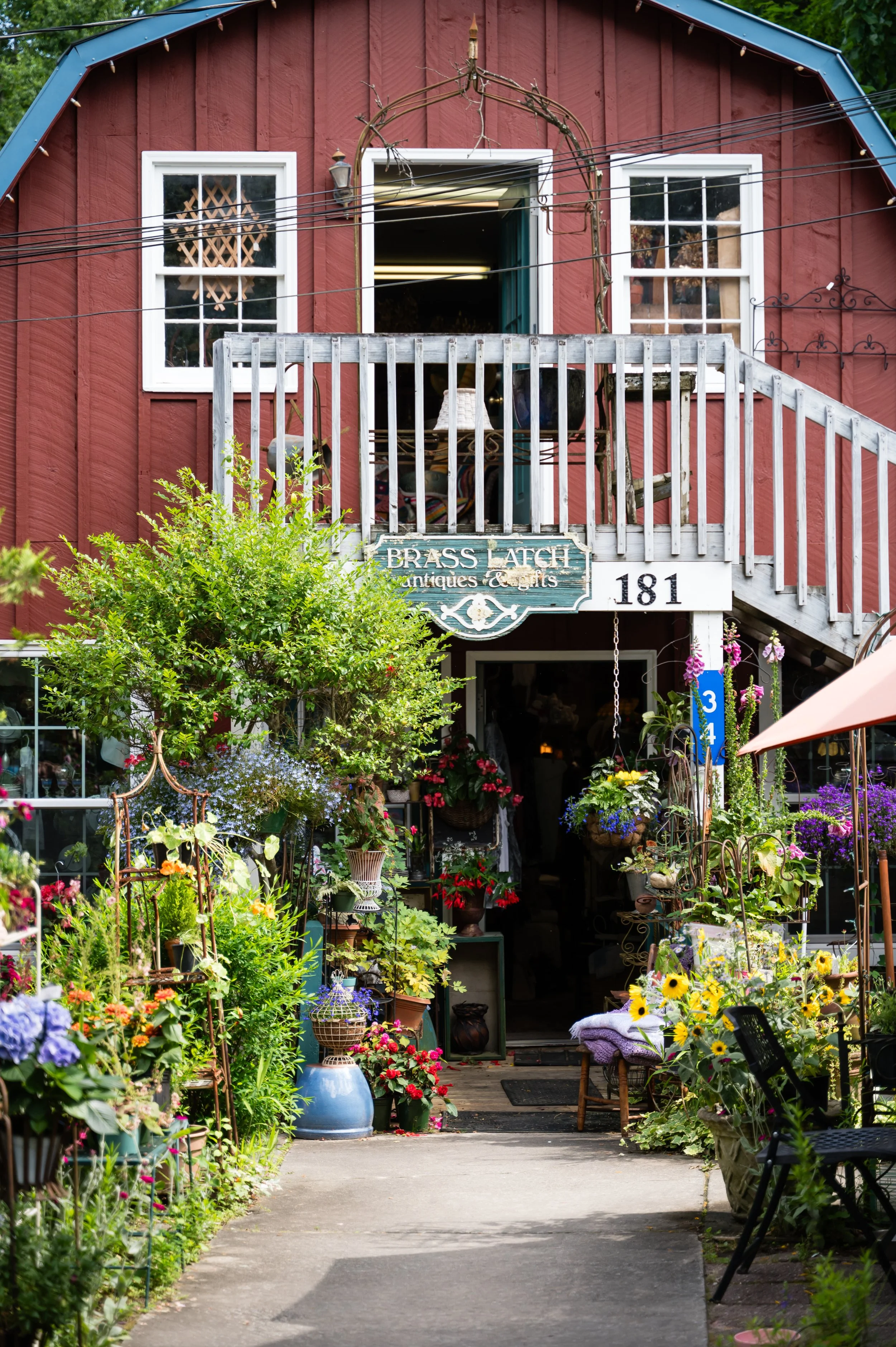 A storefront with a red wooden exterior, white-framed windows, and a sign that reads 'Brass Latch Antiques & Gifts'. The store is decorated with various plants and flowers outside, creating a garden-like entrance.
