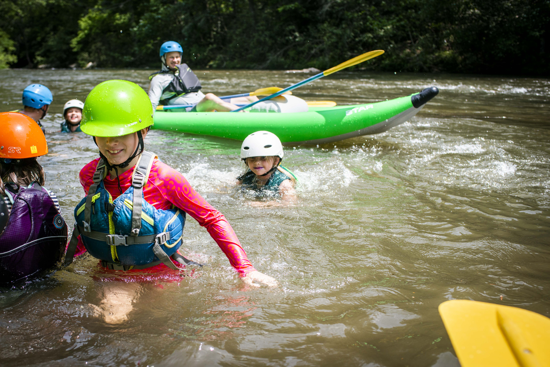 Children wearing life jackets and helmets enjoying a kayaking adventure on a river, with some children in kayaks and others in the water.