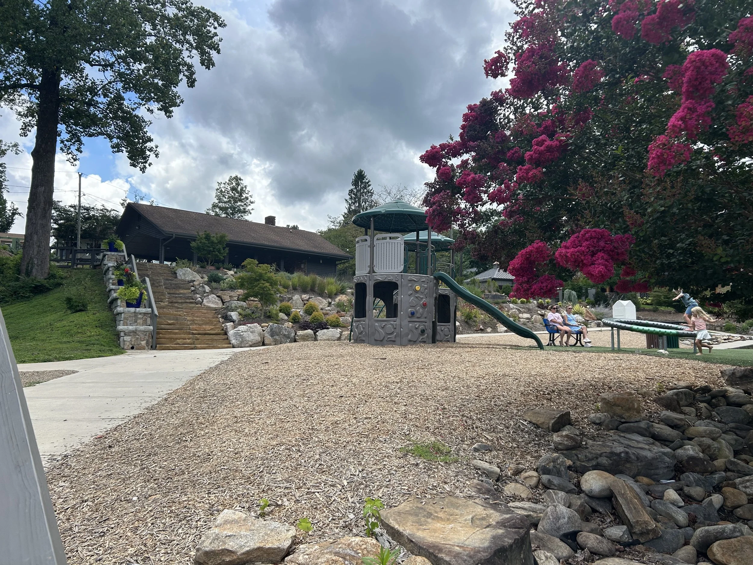 A backyard scene with a playground structure, a large pink flowering bush, a small house with stairs, and a few people sitting and children playing. There are trees and cloudy sky overhead.