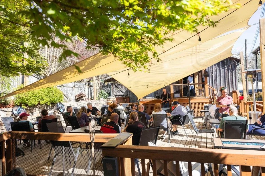 People gathered for an outdoor event or concert on a wooden deck with a yellow canopy and string lights, surrounded by trees and wooden fences.