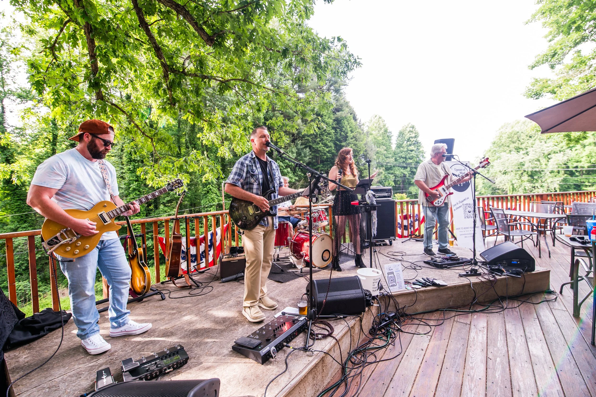 A band performing on an outdoor wooden deck, surrounded by green trees, with instruments including guitars, drums, and microphones, and various sound equipment.