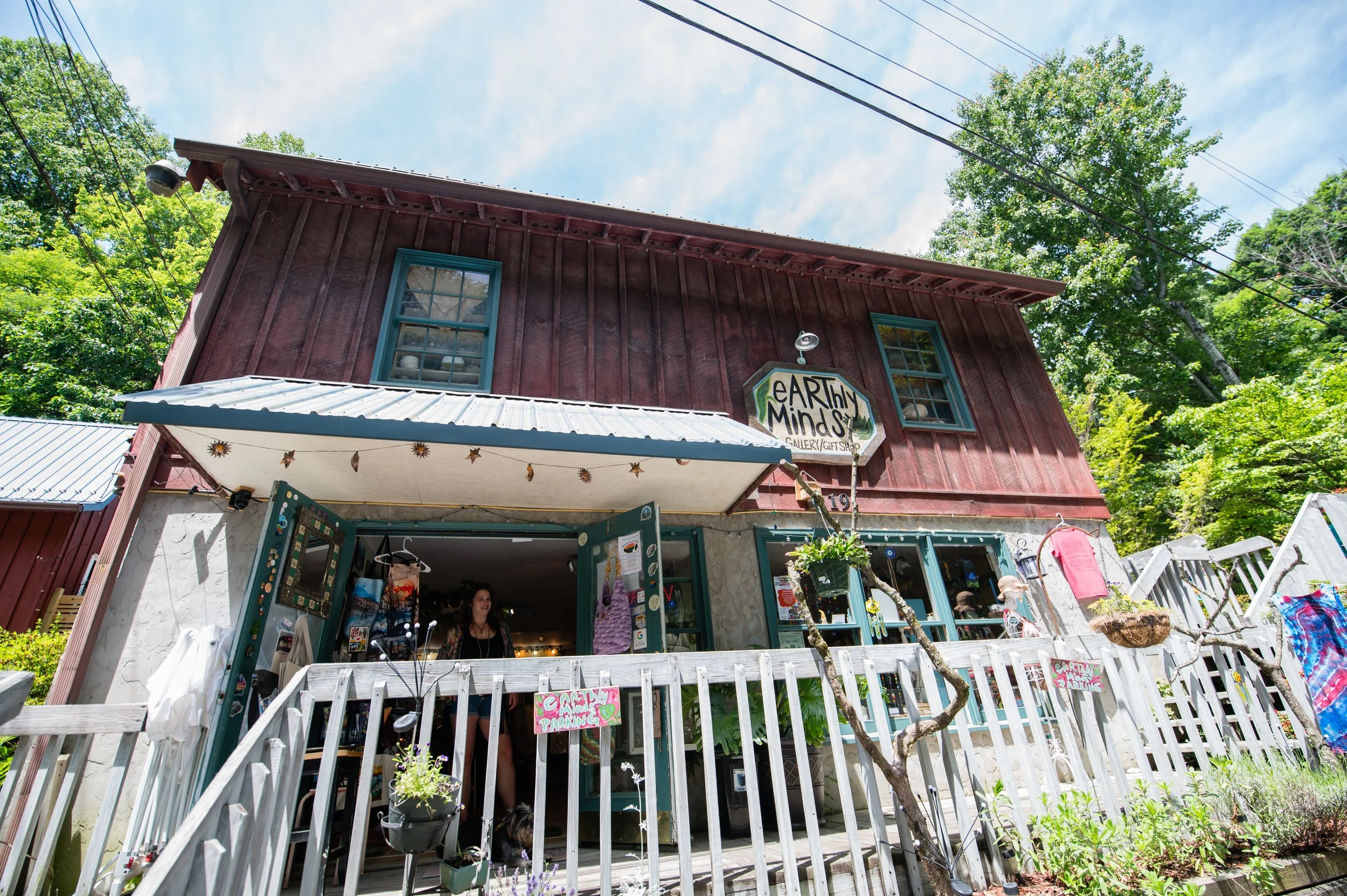 A two-story rustic red building with teal trimmed windows houses 'Earthy Minds,' an art gallery and gift shop. The building has a white corrugated metal roof with a string of decorative hanging ornaments and a front porch area with open teal doors. A