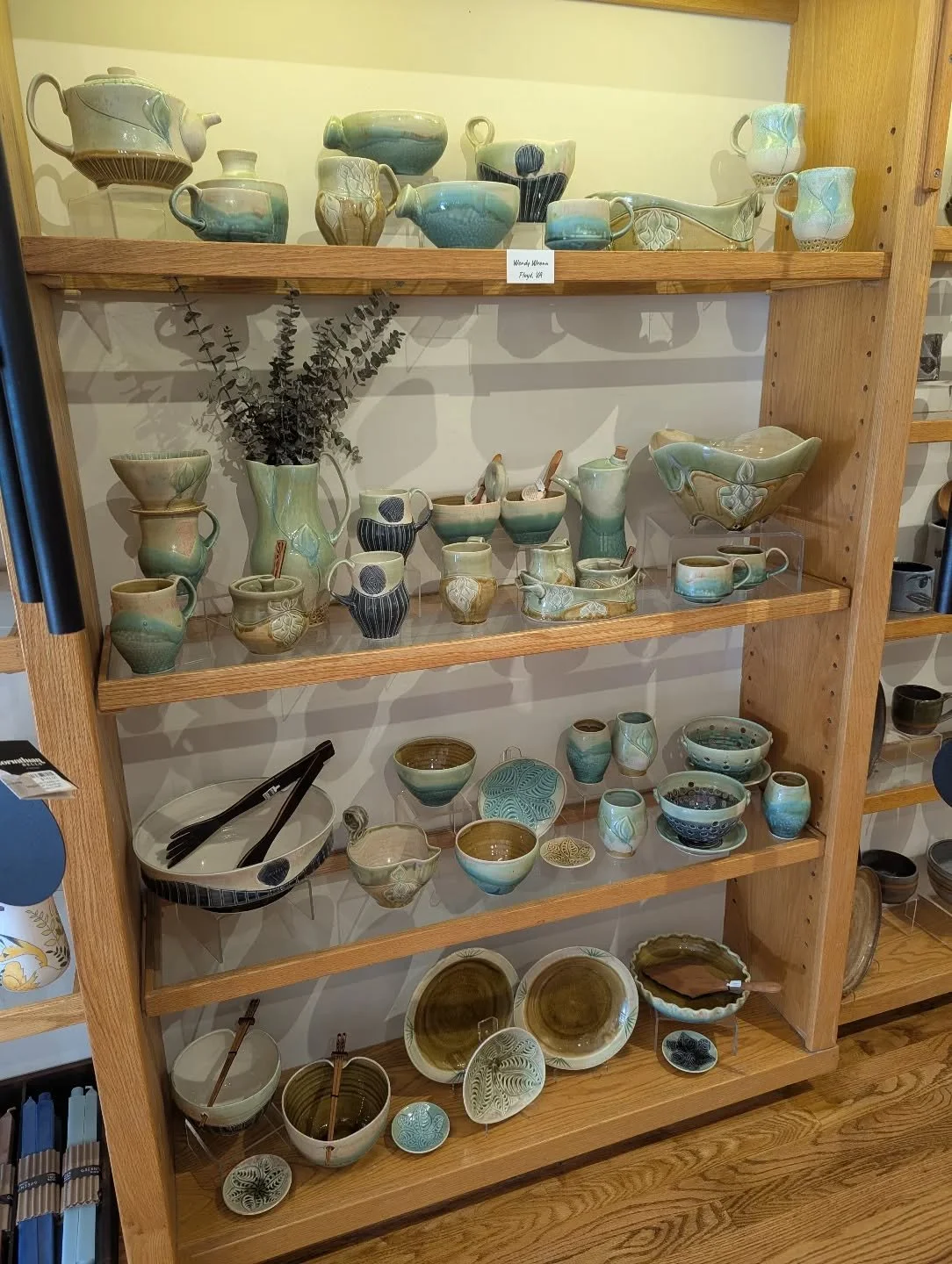 Display of various handmade pottery and ceramic dishes on a wooden shelf, including bowls, cups, and pitchers in earthy and pastel colors.