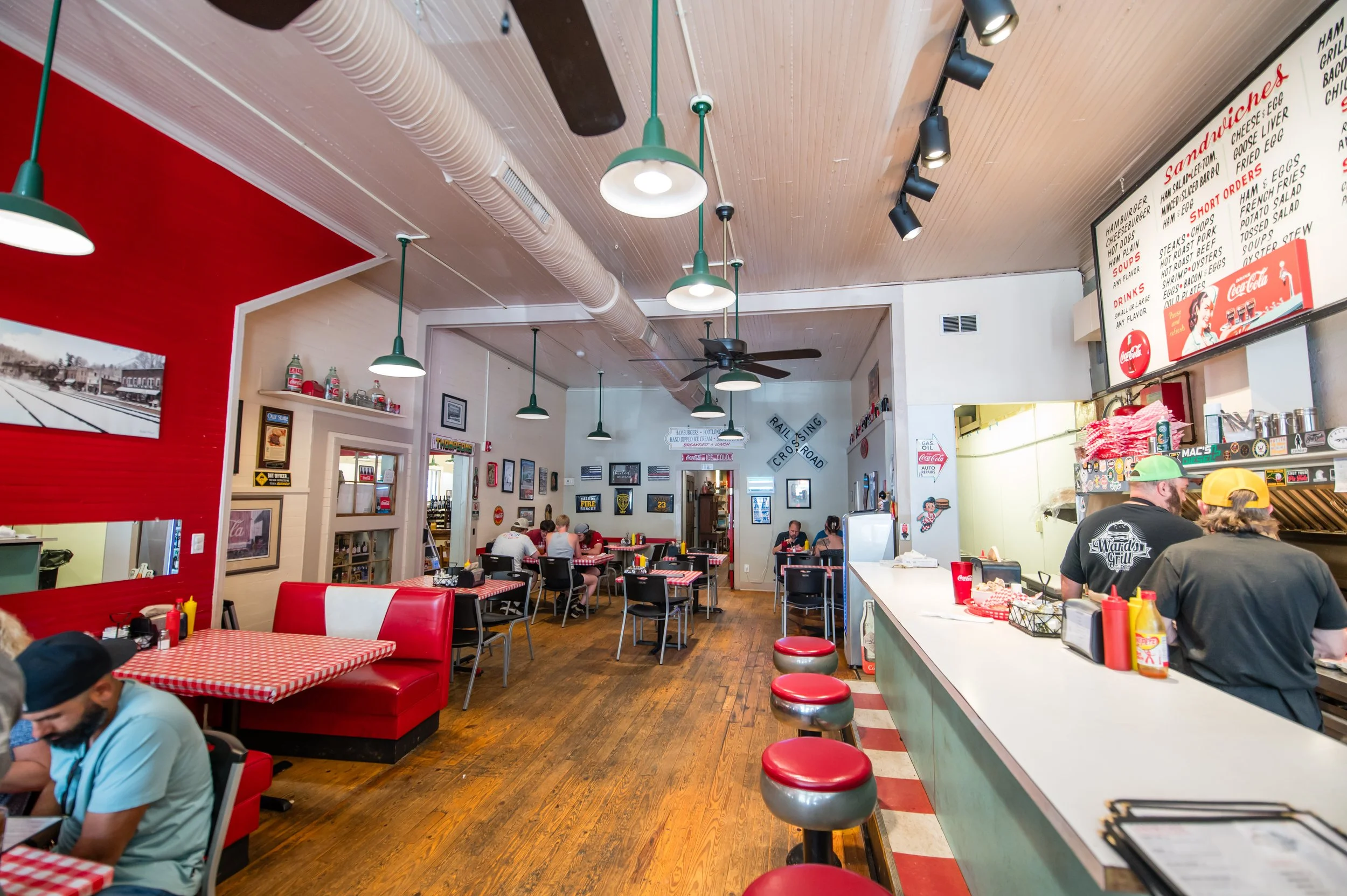 The interior of a lively American diner with red and black checkered tablecloths, red booth seating, and a counter with red stools. The ceiling has industrial-style fans and hanging pendant lights. The walls display framed photographs, vintage signs, and a menu board. Customers are seated and enjoying their meals.