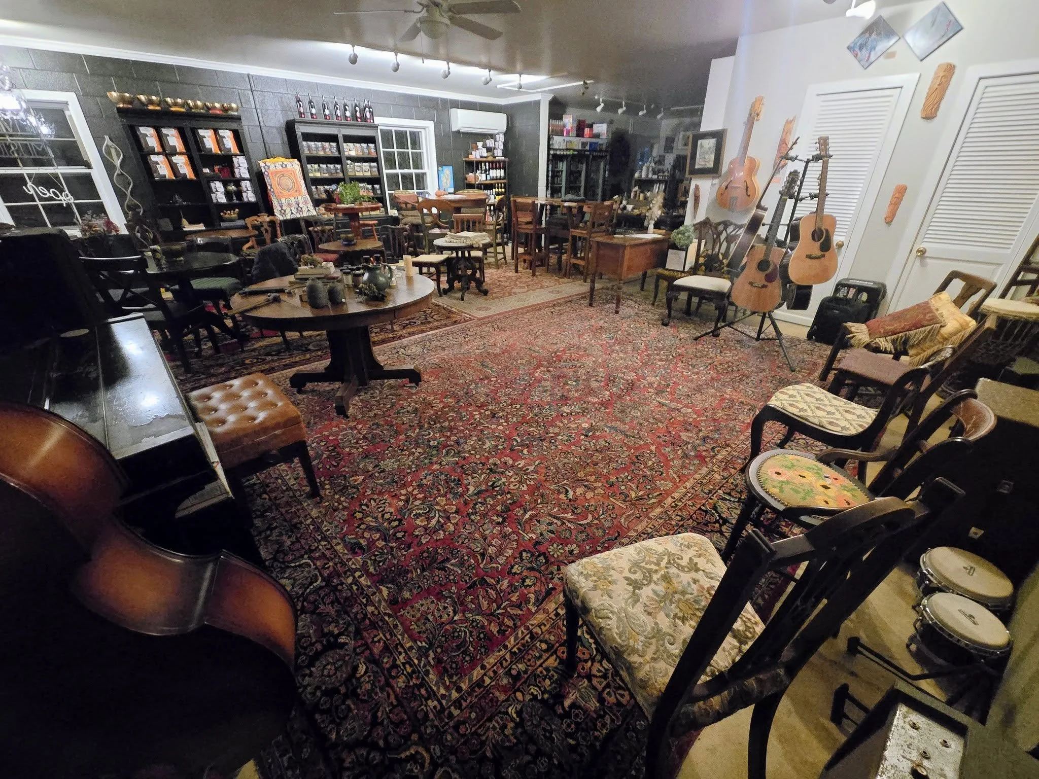 A cozy room filled with various musical instruments, chairs, and tables. The floor is covered with a large, ornate red and blue patterned rug. Guitars are hanging on the wall, and there are several chairs around the room, some with cushions. Shelves 