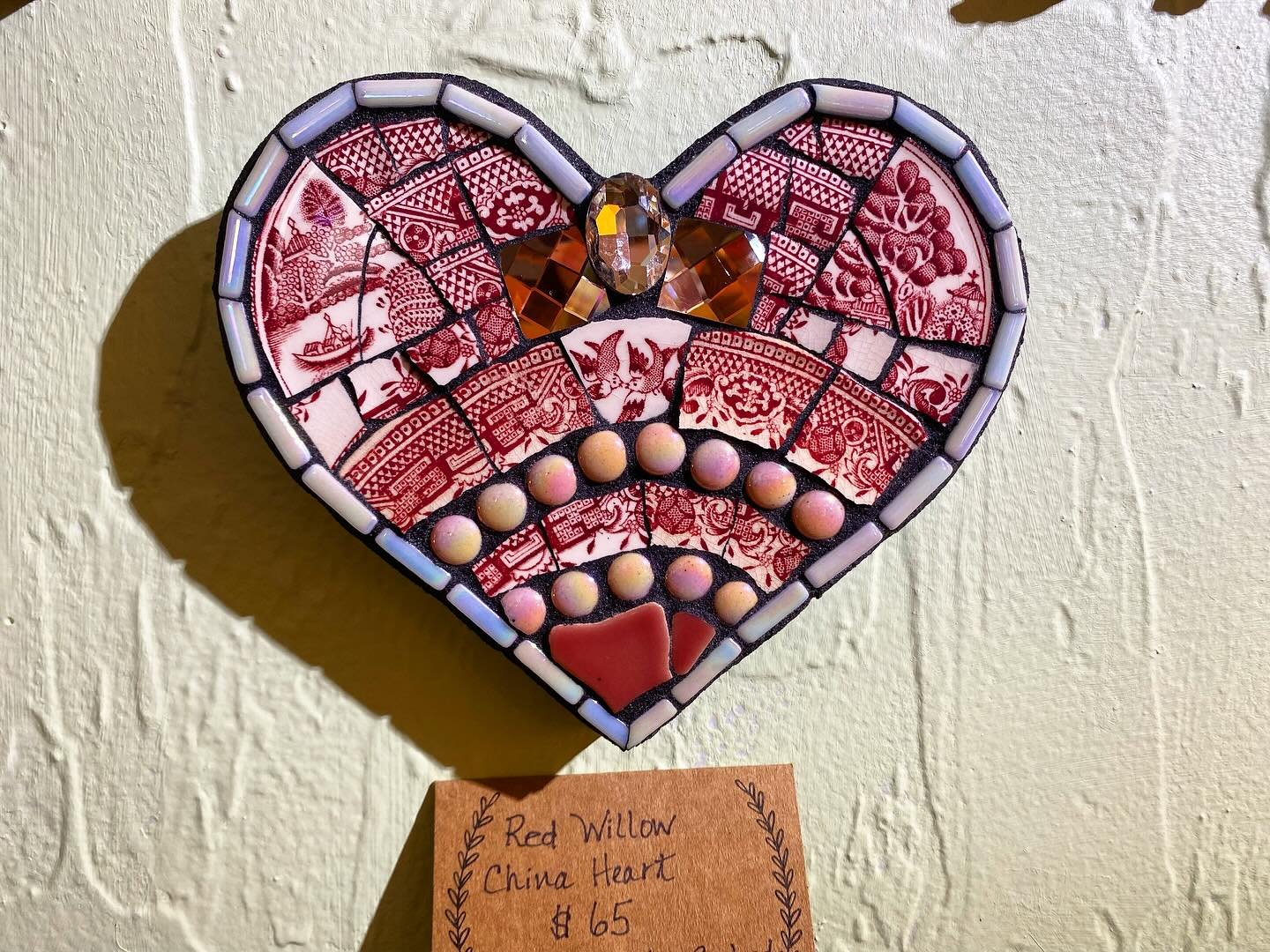 Heart-shaped mosaic artwork made of red and white paper, beads, and a gemstone, mounted on textured beige wall.