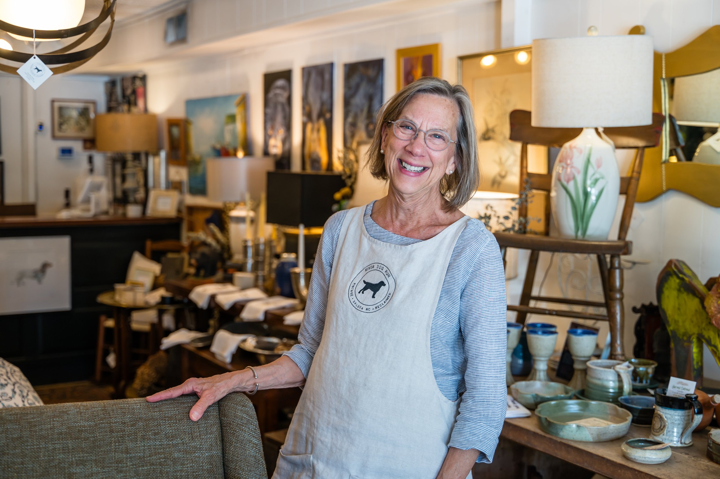 Smiling elderly woman with glasses and pearl earrings, wearing a white apron, standing inside an art gallery or craft shop with paintings and pottery on display.