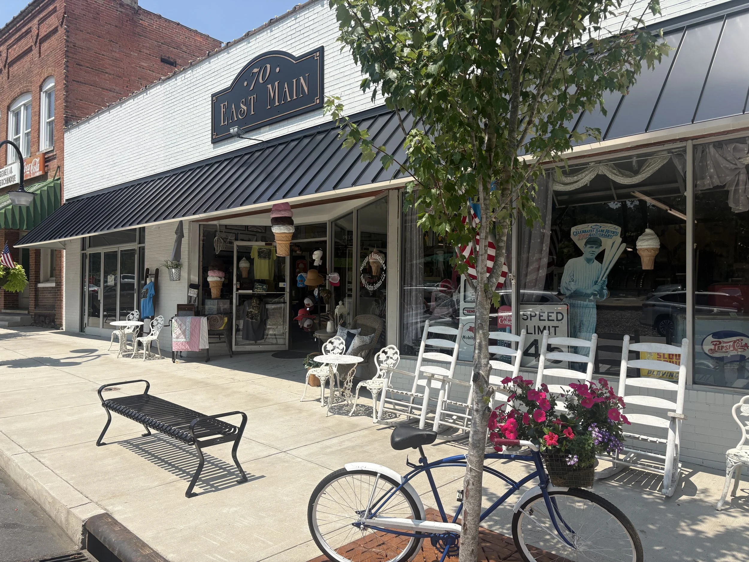 A storefront with a black sign reading 70 East Main, large windows displaying various items inside, a small outdoor seating area with white metal chairs and tables, and a bicycle parked near a small tree with pink flowers planted in a basket. The sid