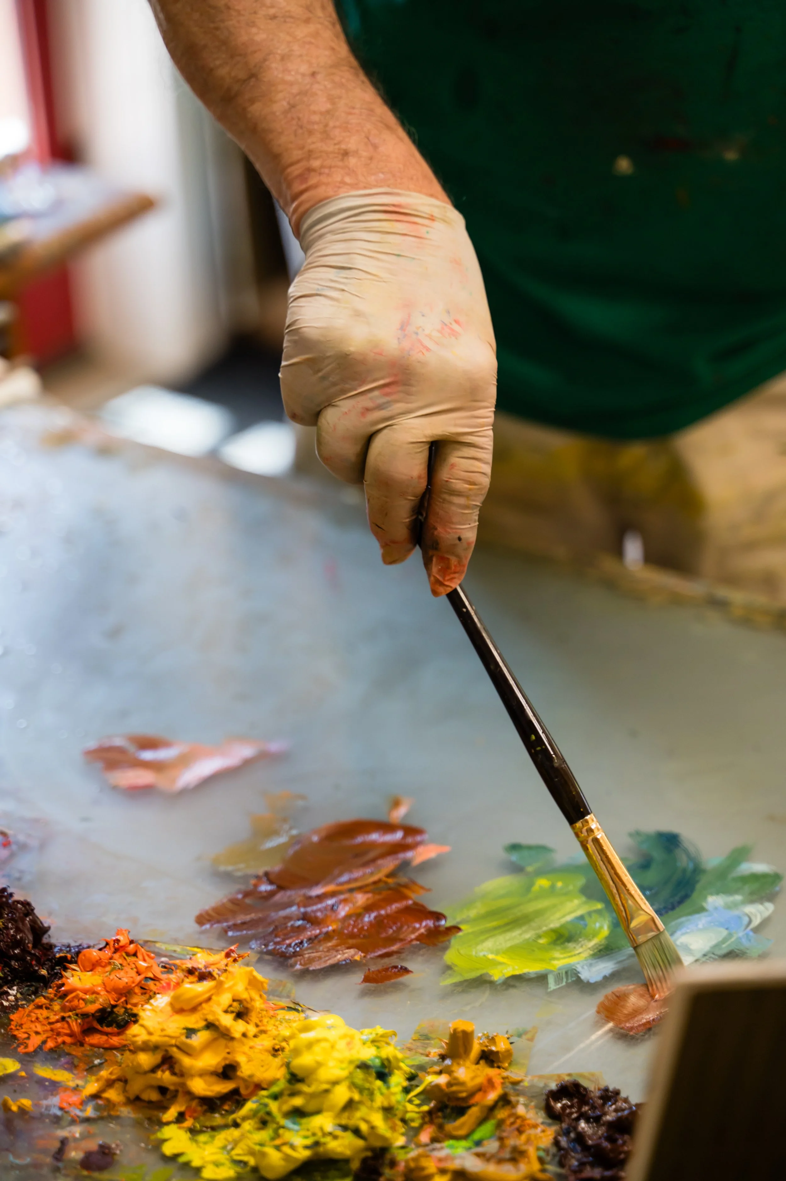 Close-up of a person's hand holding a paintbrush, mixing bright colors of paint on a palette. The background shows a blurred workspace with additional colors and art supplies.