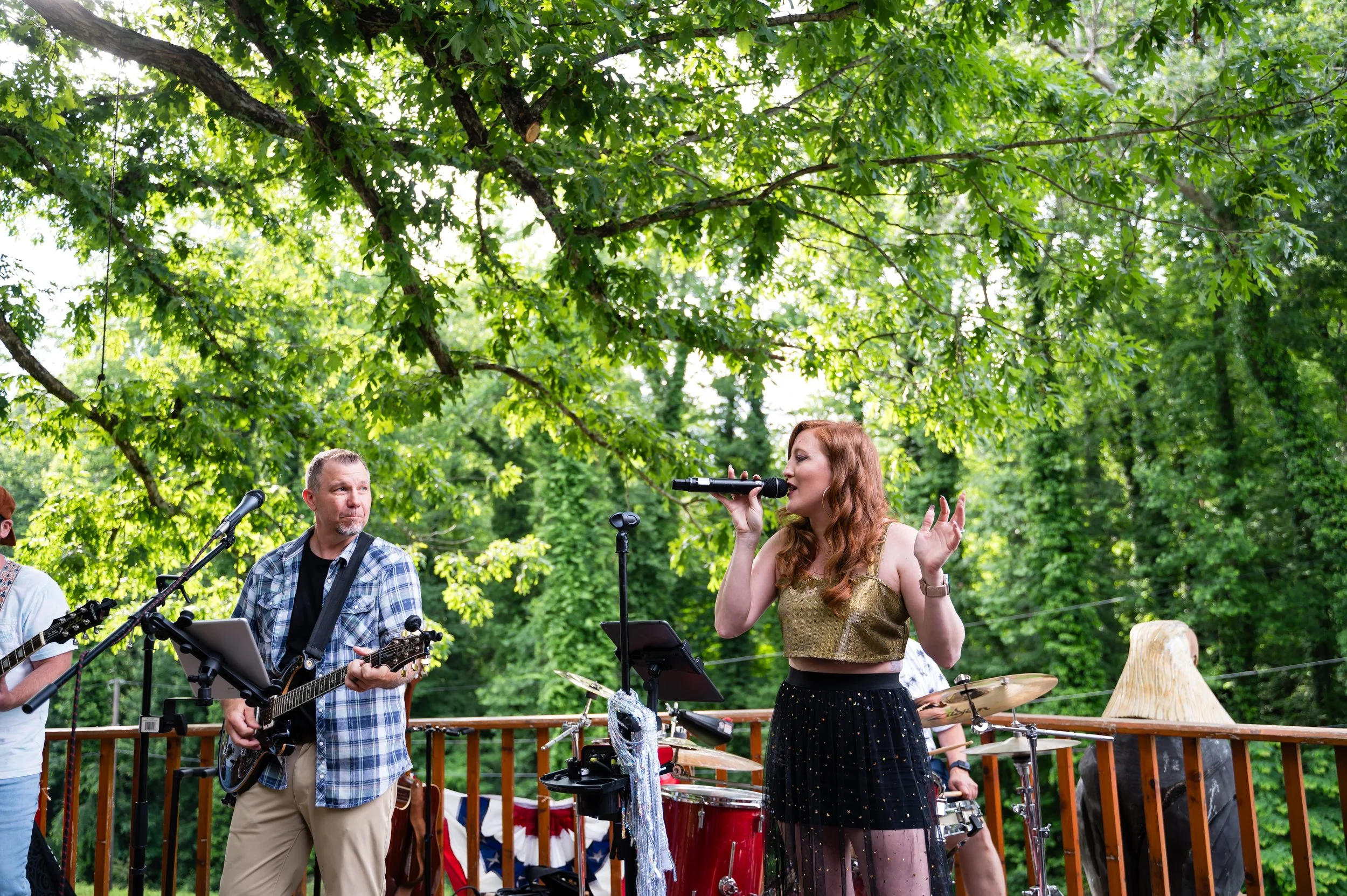 A group of musicians performing outdoors under a large leafy green tree, including a woman singing into a microphone and another man playing guitar.