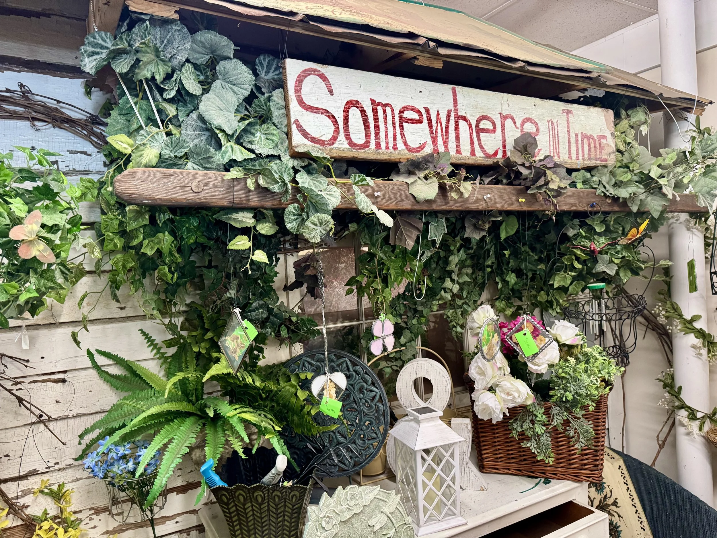 Decorative display of garden and home decor items, including green plants, artificial flowers, wicker baskets, decorative tags, and garden ornaments arranged on shelves.