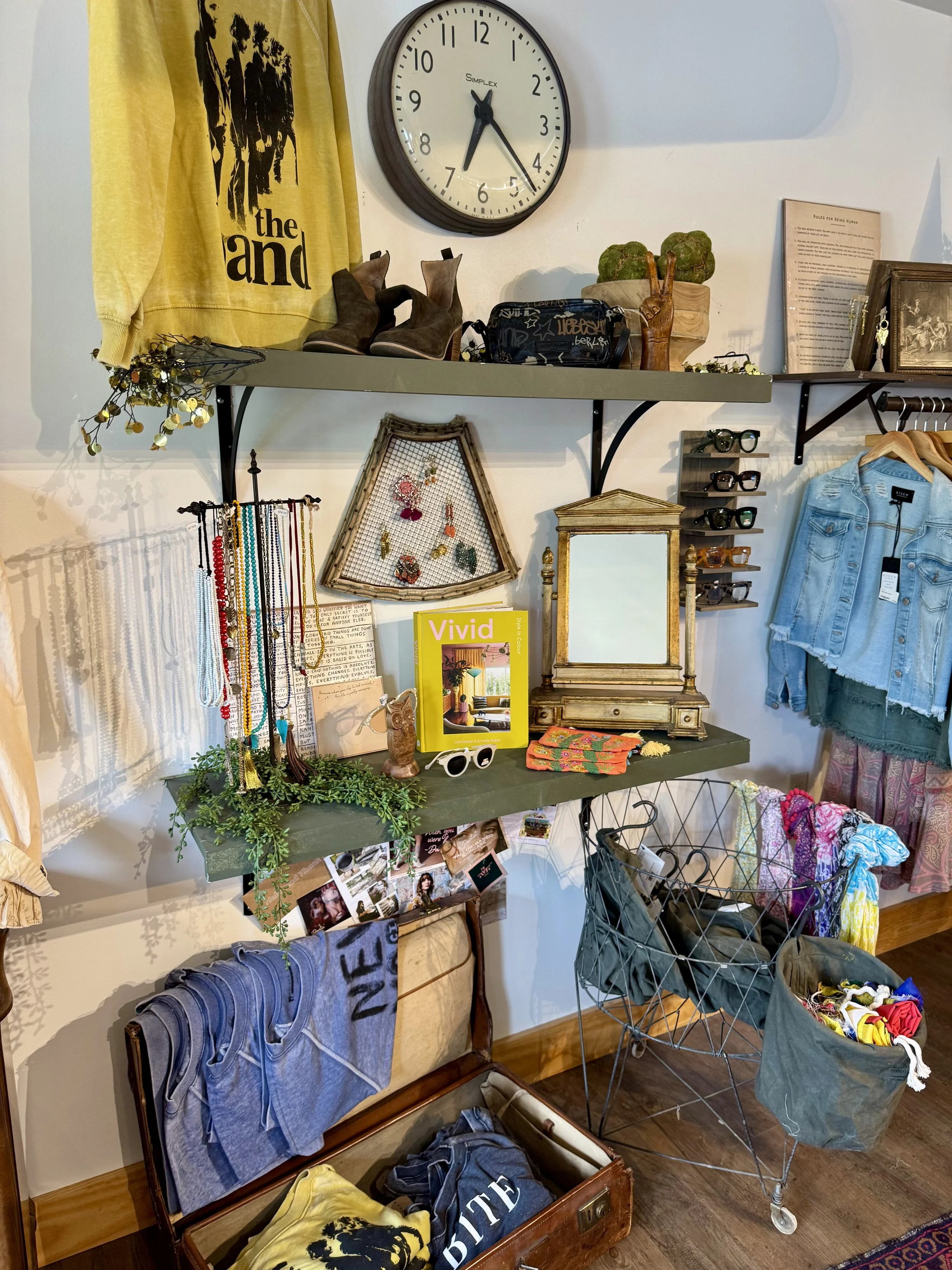 A retail display with a wall shelf holding shoes, jewelry, and decorative items. A yellow sweatshirt hangs nearby, and a wall clock shows 7:29. Below the shelf are racks with sunglasses, a vintage dresser with jewelry, and a shopping cart filled with