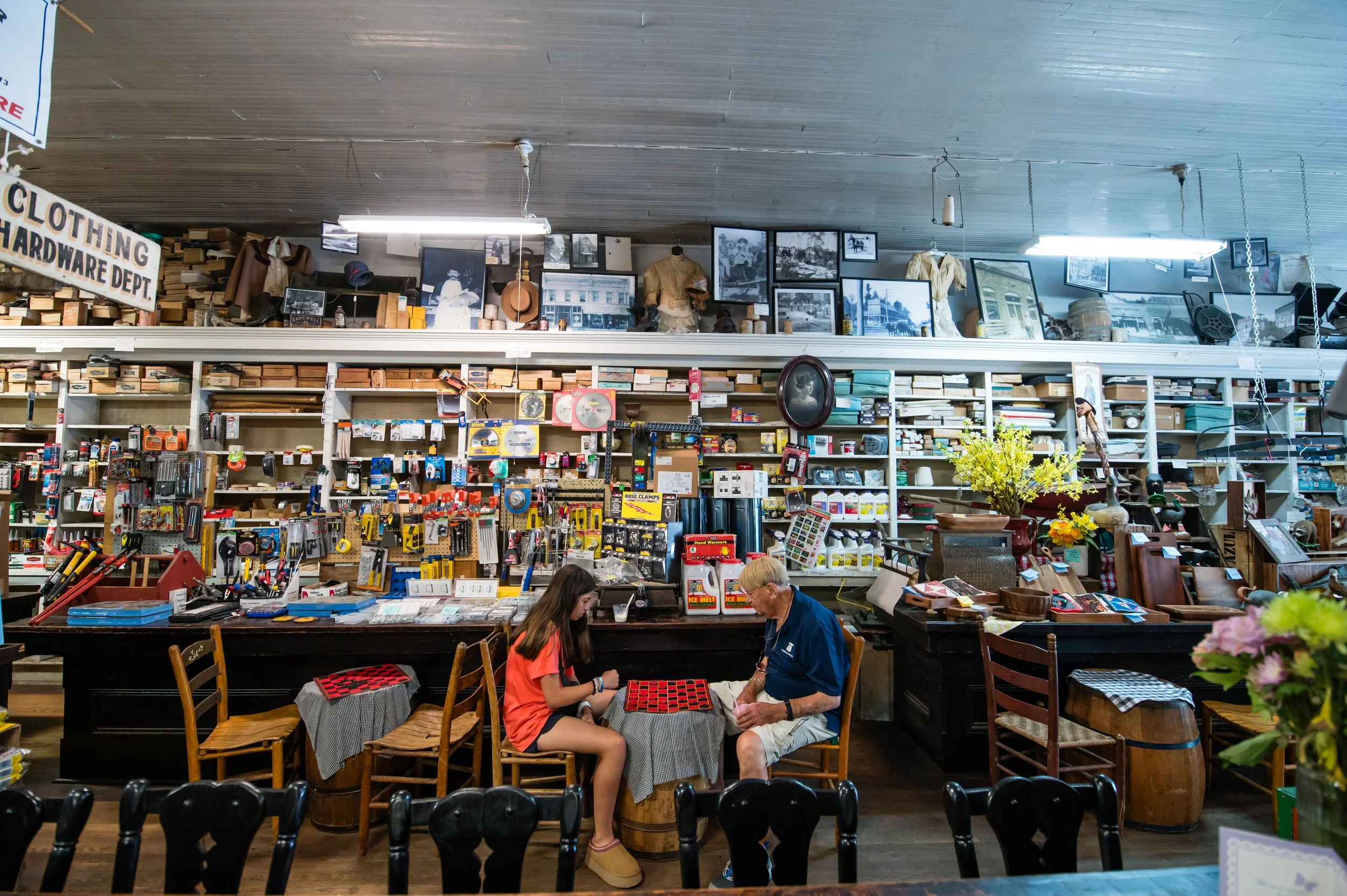 People playing checkers at a table inside a vintage store filled with pictures, furniture, and hardware items.