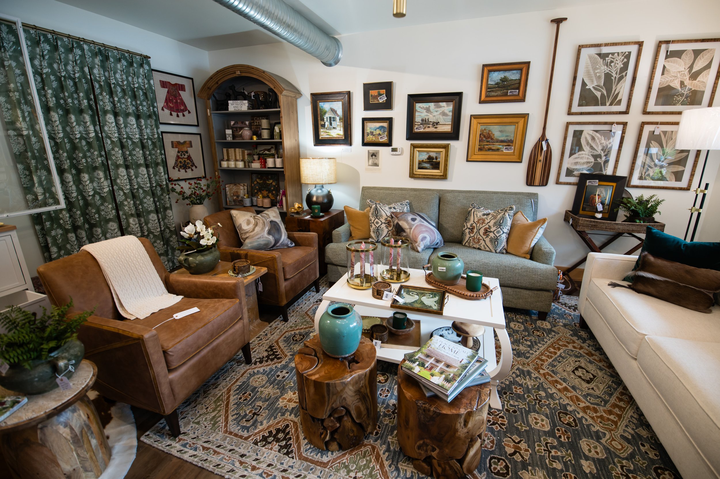 Living room decorated with artwork, plants, and furniture including sofas, armchair, and a coffee table with decorative items and magazines.