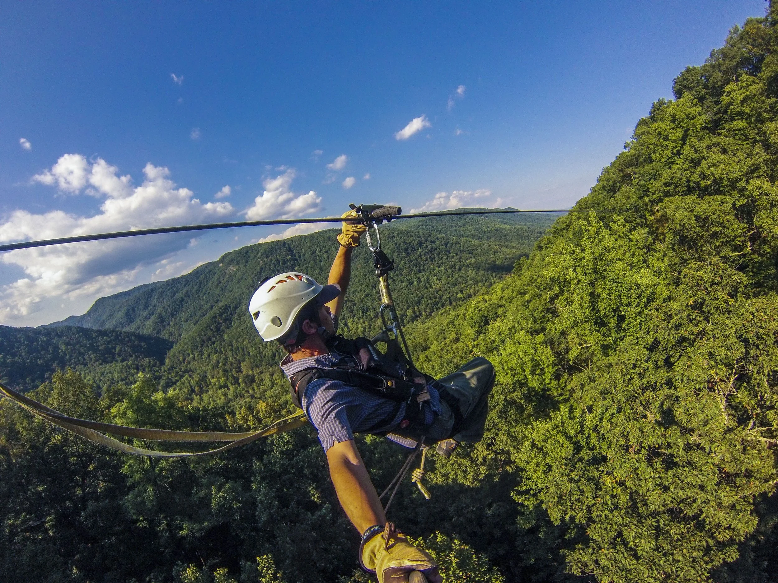 Person wearing a white helmet and yellow gloves stabilizing a zip line in a green mountainous area on a sunny day.