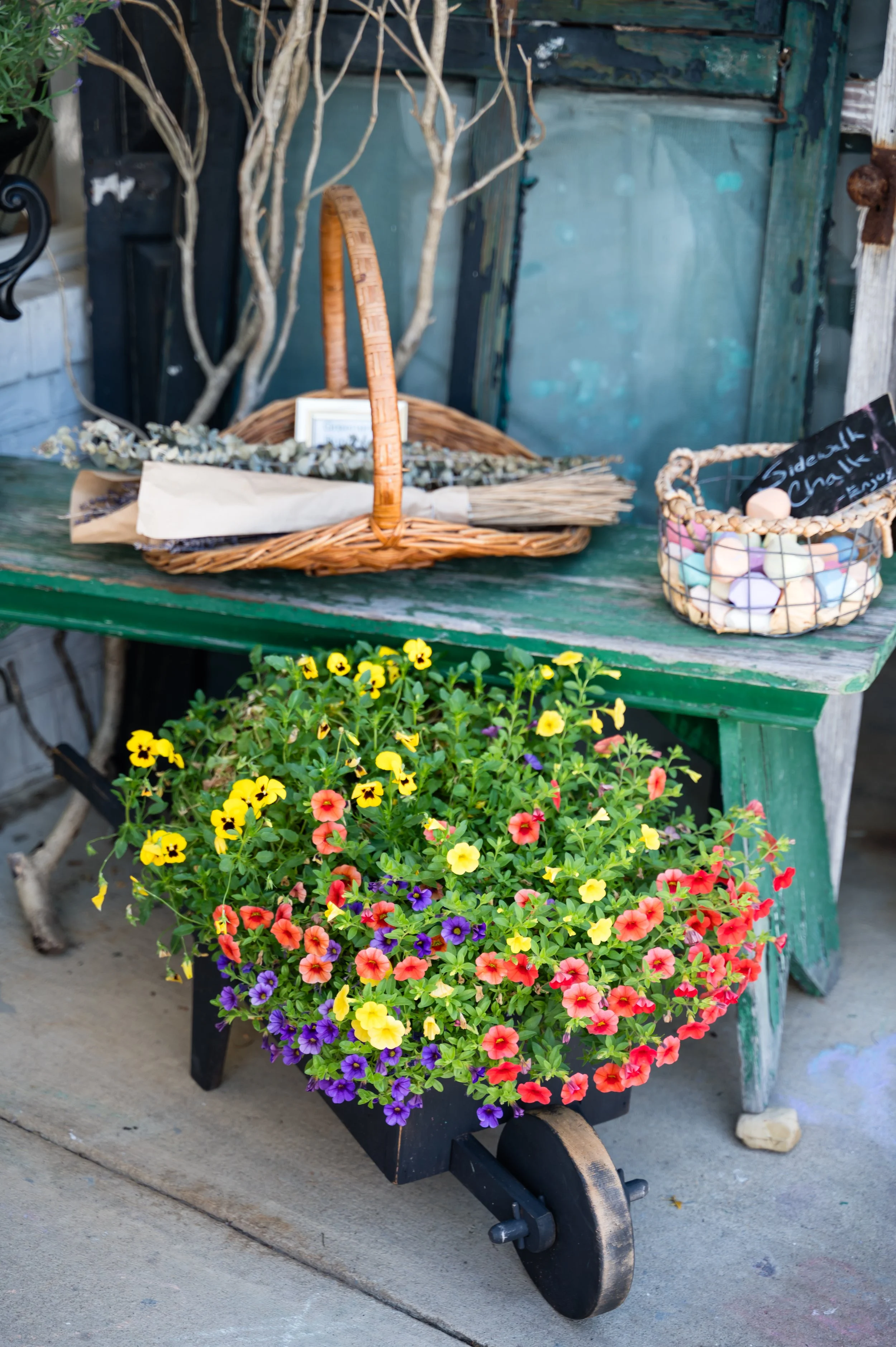 A black wooden cart with flowers, a basket of pastel-colored candies, and dried lavender. The cart is outdoors on a concrete surface, with a large colorful flower pot and yellow blossoming plants visible.