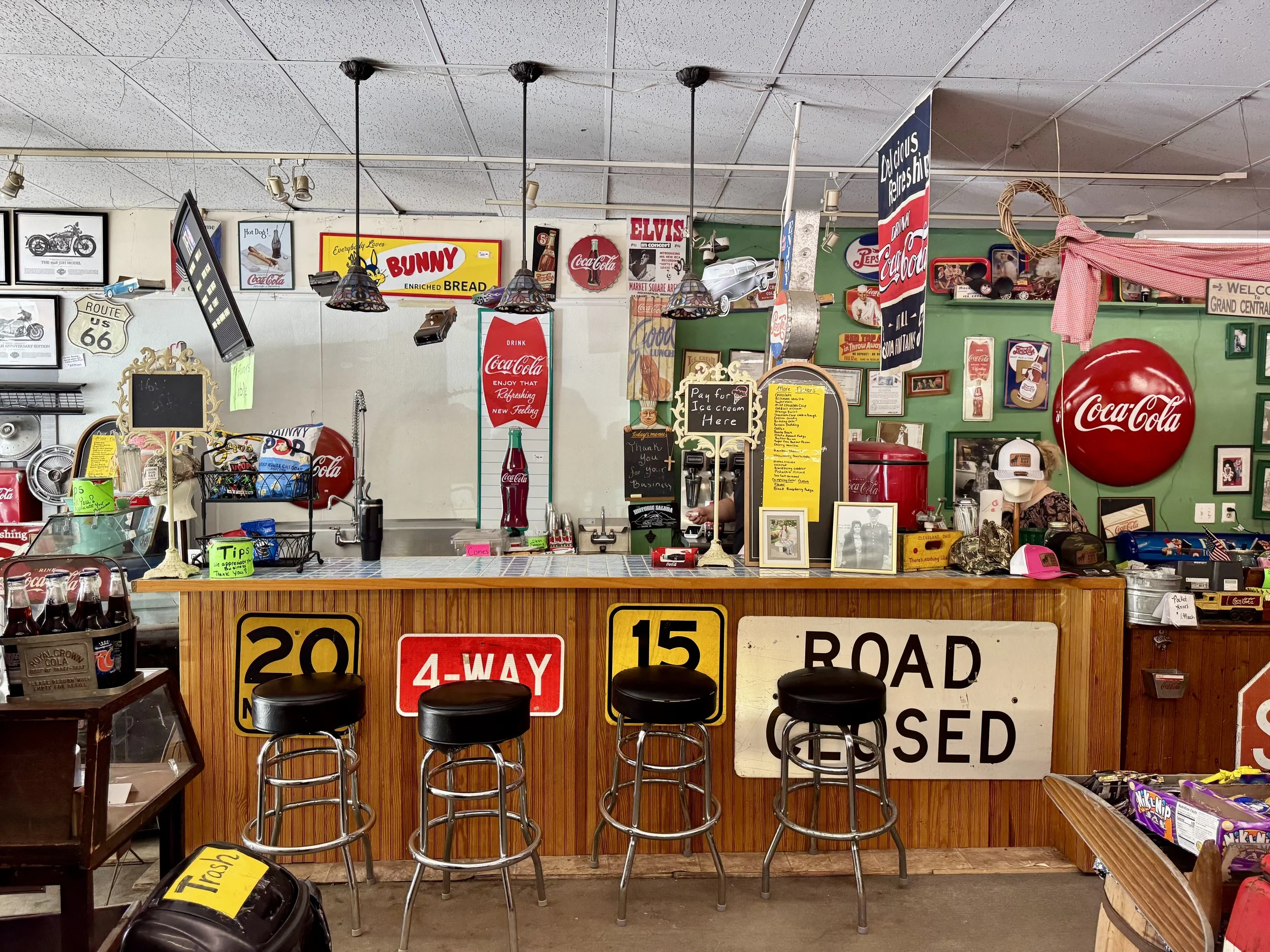 Inside a vintage-style diner or ice cream shop with memorabilia and signs, including Coca-Cola advertising, Route 66 sign, T. Rex model, and various decorative items. There are barstools at a counter with signs for 20 and 15 mph speed limits, and a c