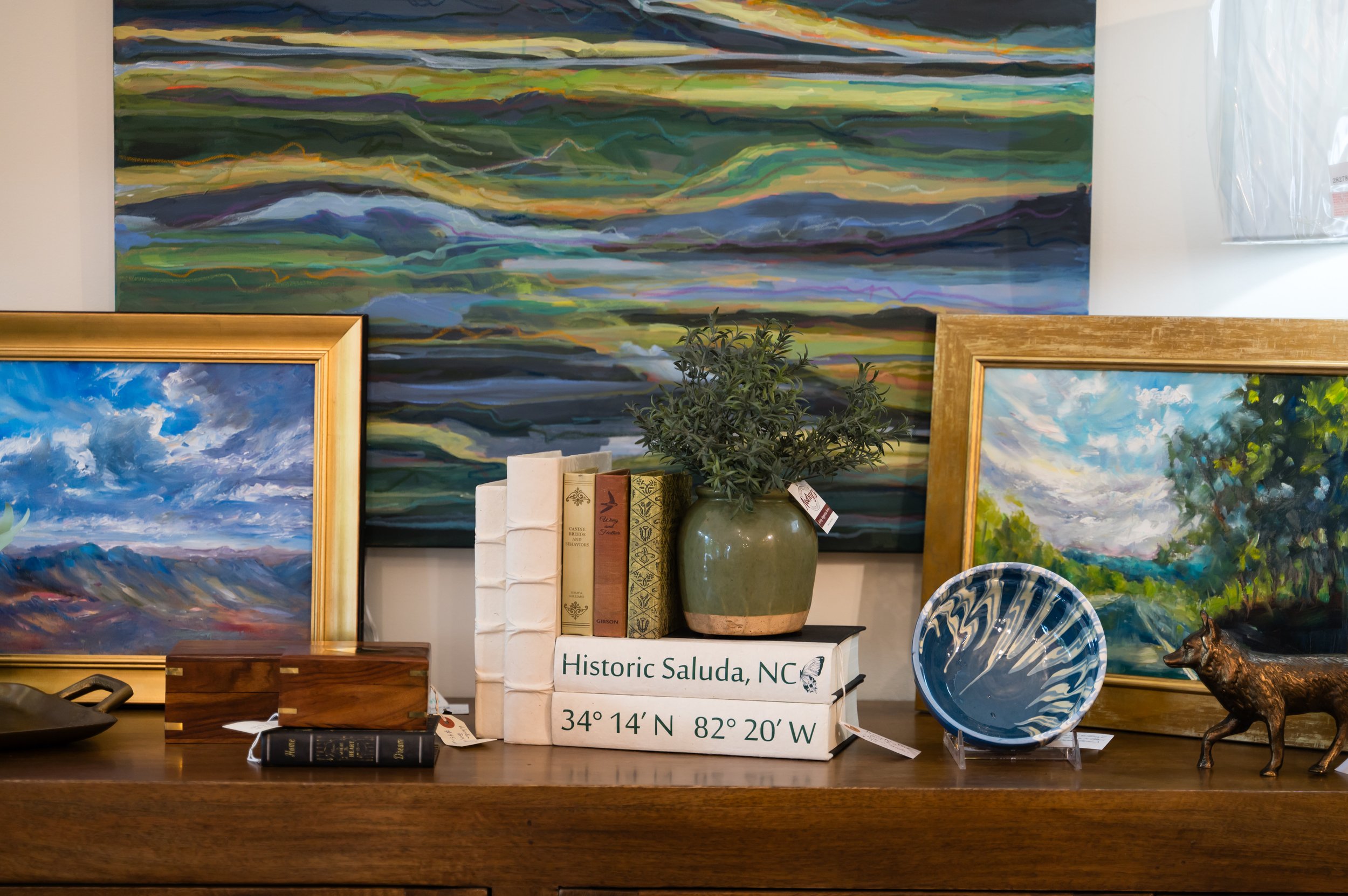 Various landscape and seascape paintings along with decorative items and books displayed on a wooden surface, with a large abstract painting on the wall behind them.