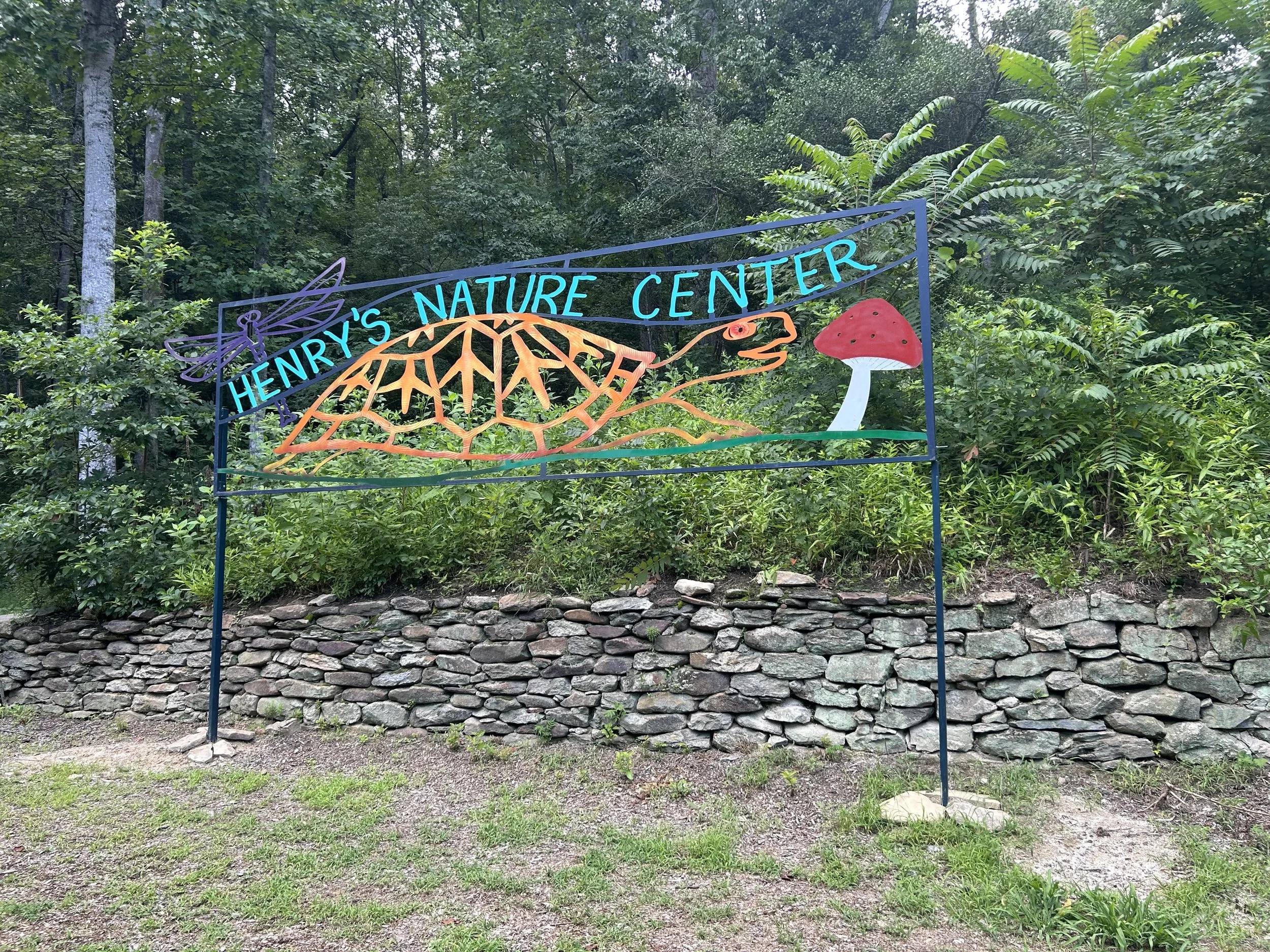 Colorful metal sign for Henry's Nature Center featuring a dragonfly, a topographical map, a lizard, and a mushroom, set outdoors with trees and a stone wall in the background.