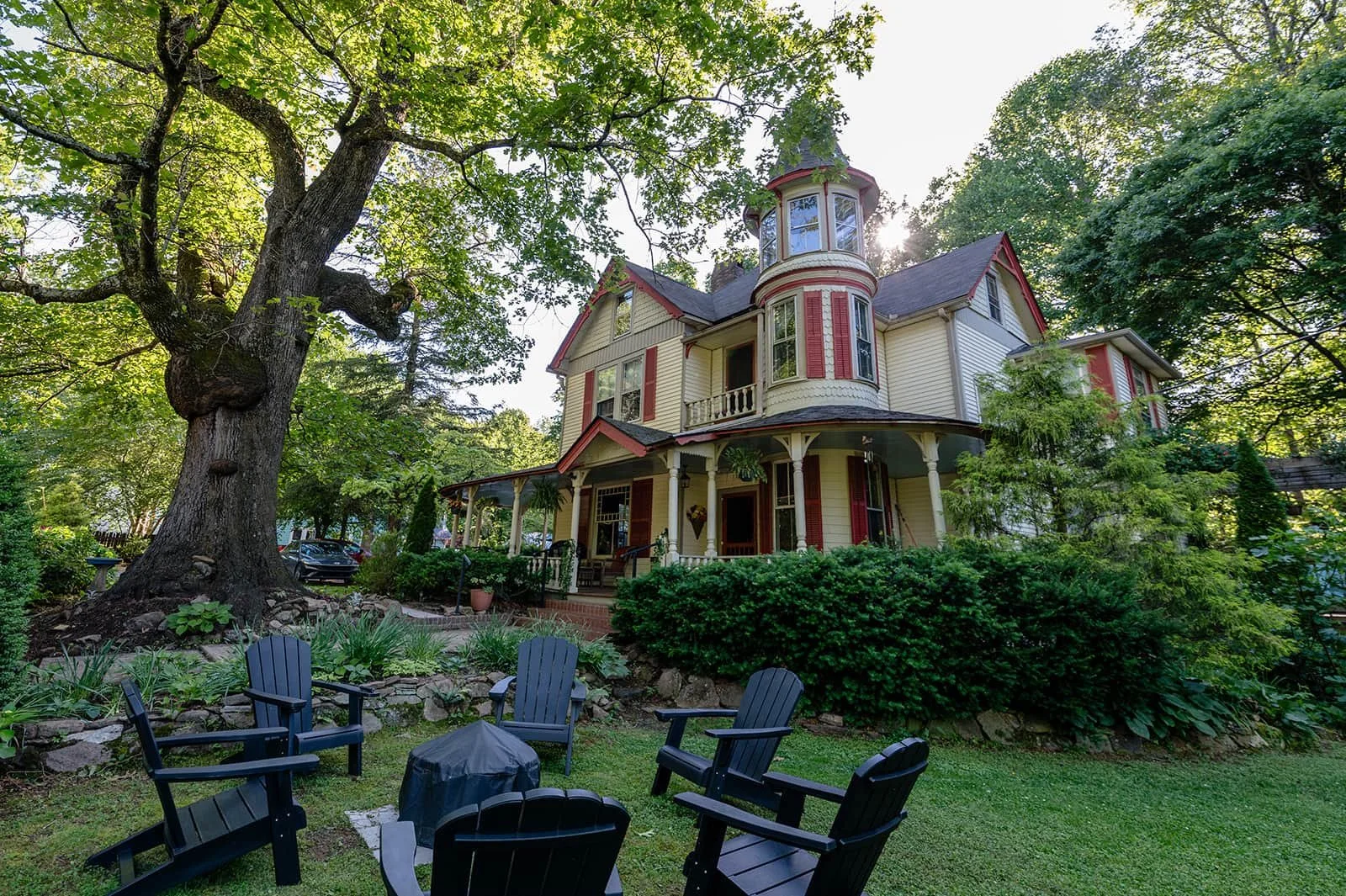 A large, Victorian-style house with a turret, cream-colored siding, red accents, and a wrap-around porch, surrounded by lush green trees and shrubbery. In the foreground, there are several black Adirondack chairs and a small table on a grassy yard.