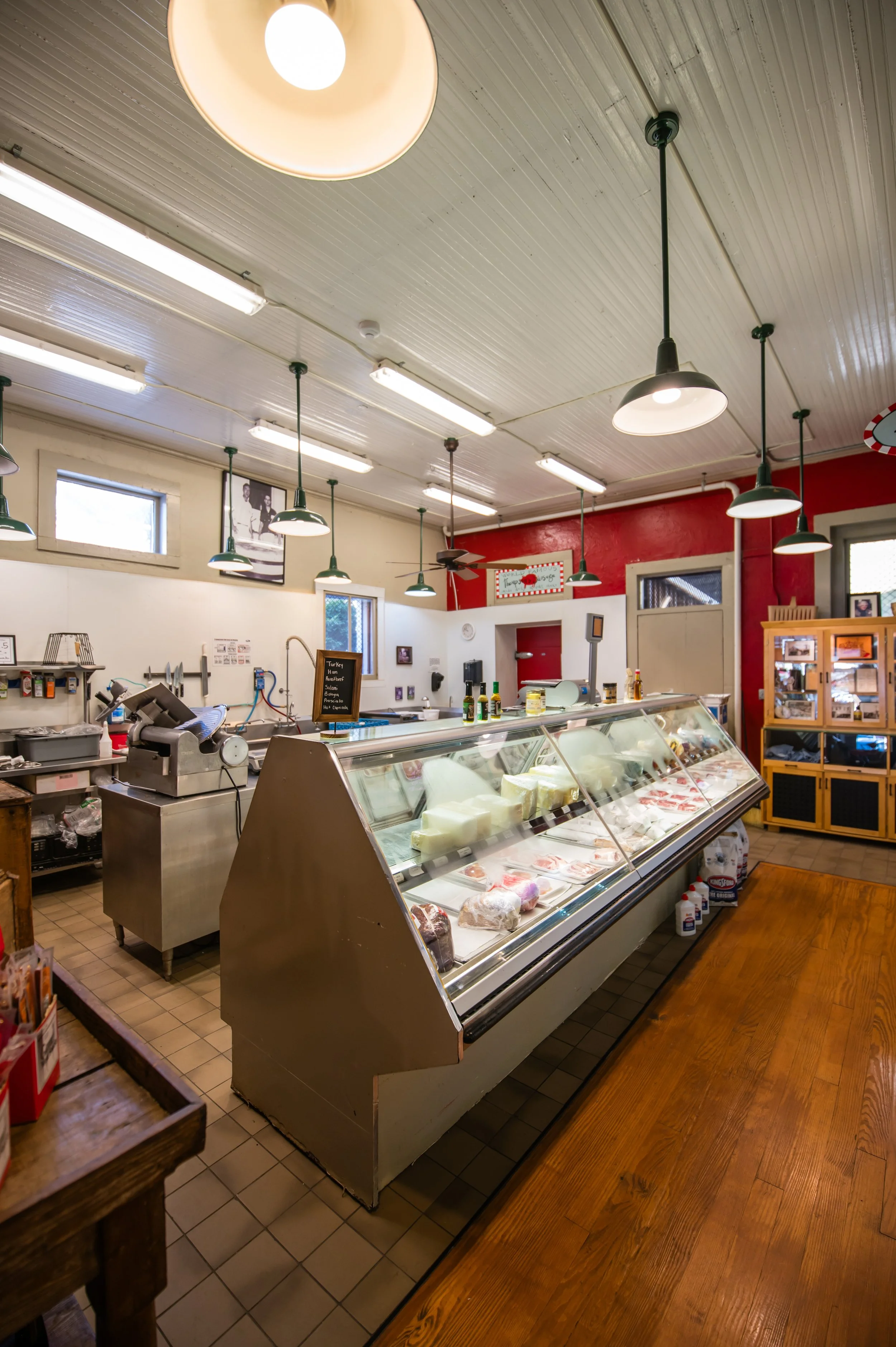 Inside a grocery store or deli with a glass display case full of meats and cheeses, overhead lighting, and a red wall in the background.