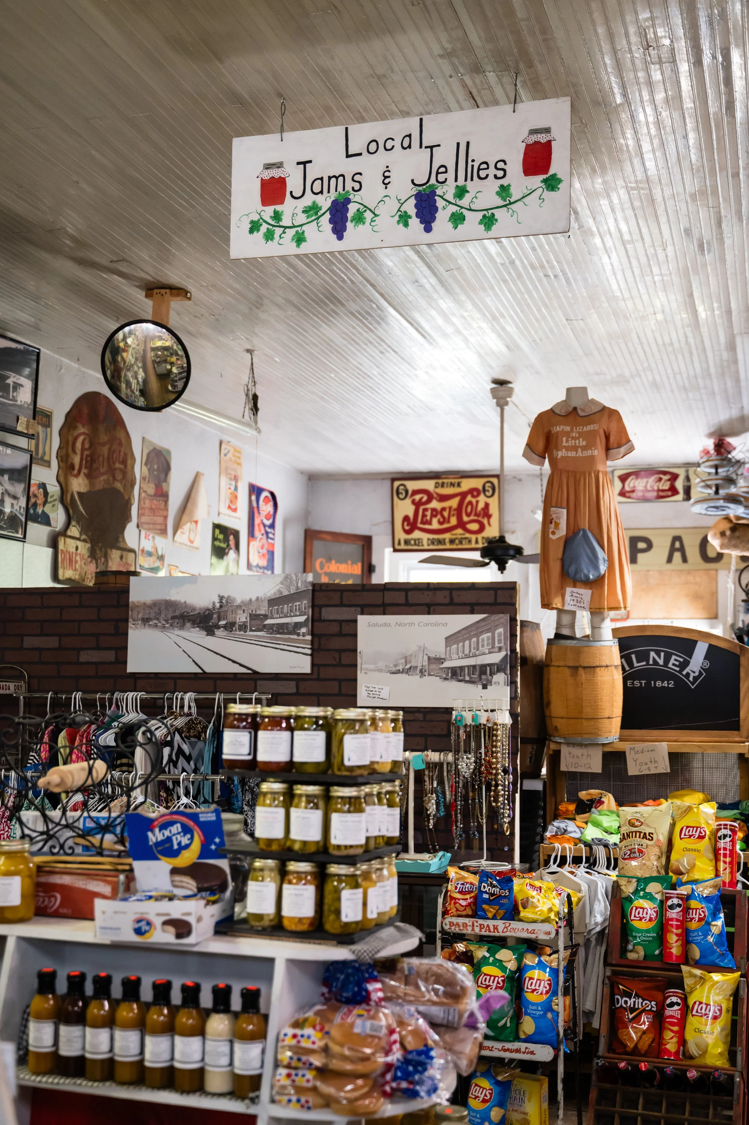 Inside a store with vintage decor, including a sign reading 'Local Jams & Jellies,' shelves with jars of jam, various snacks, bottled drinks, and vintage advertisements on the wall.