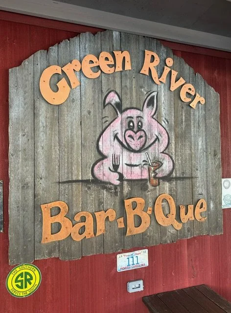 Sign for Green River Bar-B-Que with a pink pig holding a fork and a drink, mounted on a rustic wooden background.