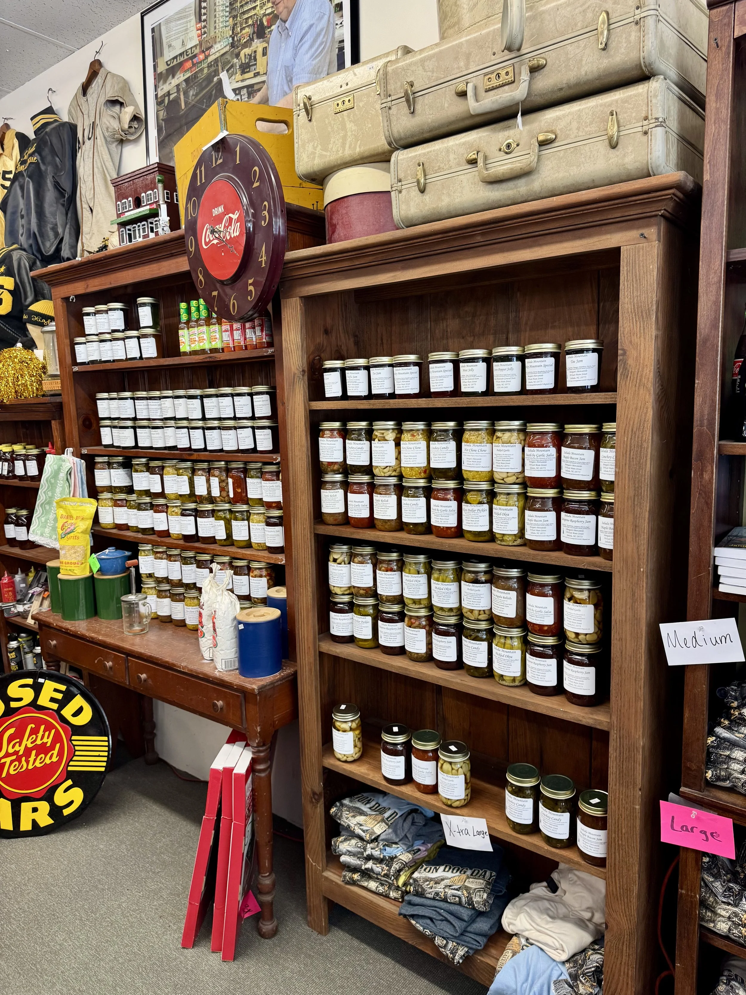 A wooden shelf displaying multiple jars of homemade jam, labeled with various flavors. There are cloth items folded on the bottom shelf, with handwritten signs indicating sizes like 'Xtra Large' and 'Large' nearby. The scene appears to be in a store 