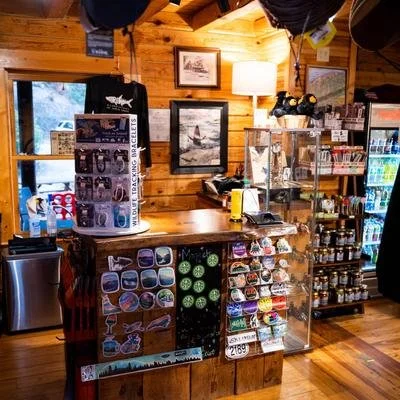Interior of a wooden shop with beverages, snacks, and souvenirs displayed on shelves and a counter, with a framed picture on the wall and a window showing an outdoor scene.