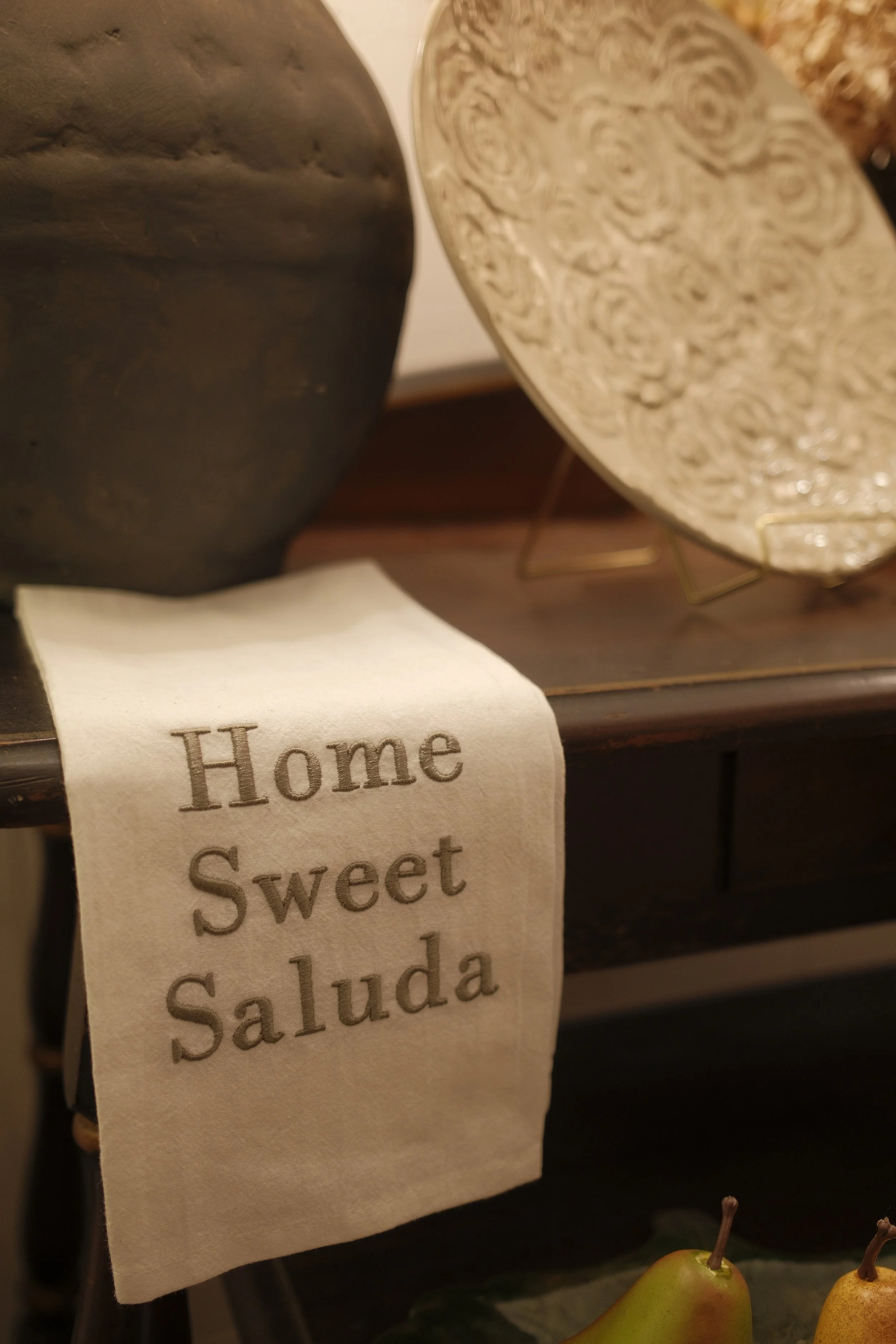 Close-up of a cream-colored dish towel with the words 'Home Sweet Saluda' embroidered in brown, hanging over a dark surface. Part of a pear is visible in the bottom right corner.