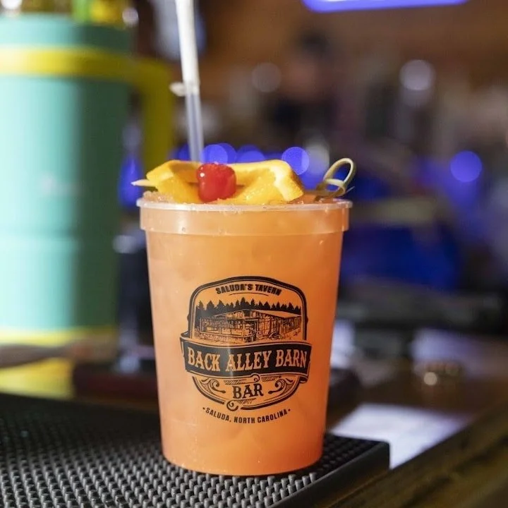 A tropical cocktail with a straw, garnished with a slice of pineapple and a cherry, on a bar counter at Back Alley Barn Bar in Saluda, North Carolina.