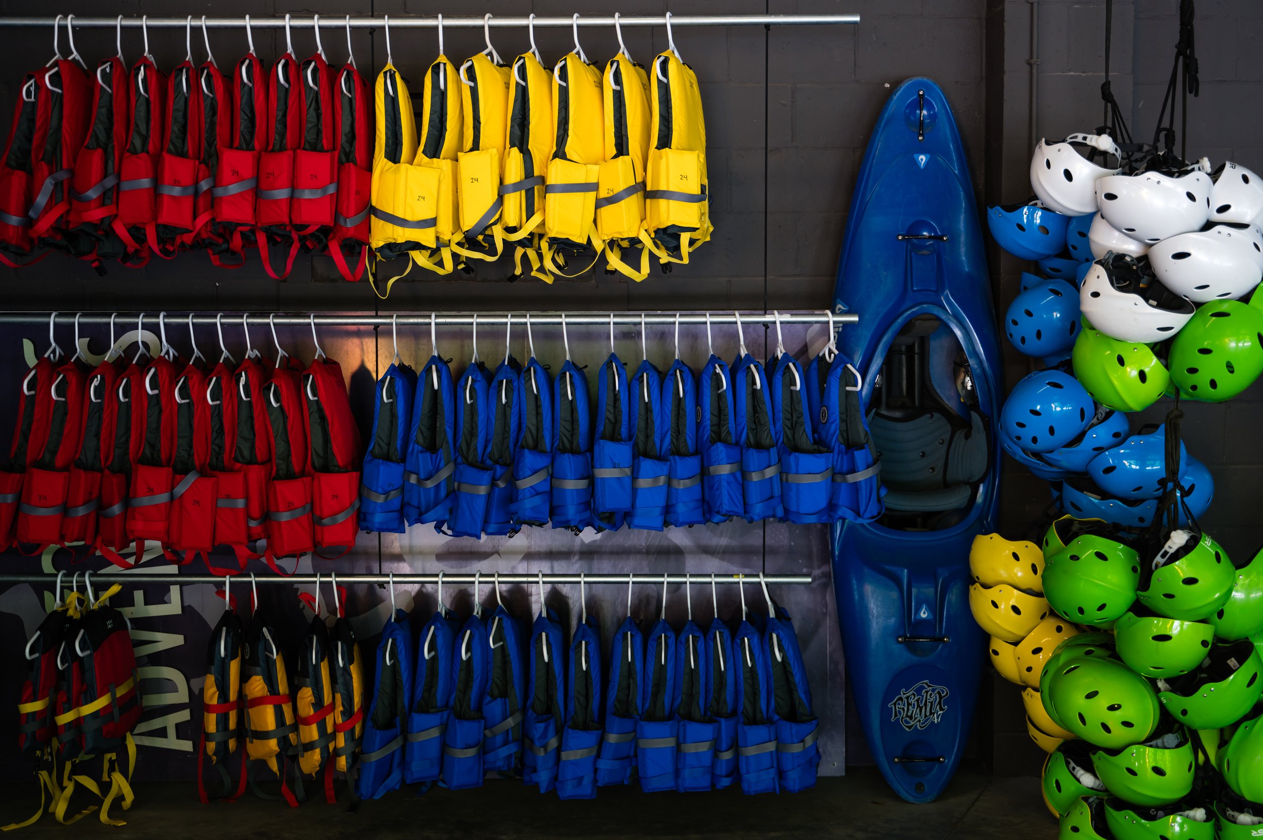 Display of life jackets in red, yellow, and blue hanging on racks with helmets and a sled on the side, likely in a sporting goods or safety gear store.
