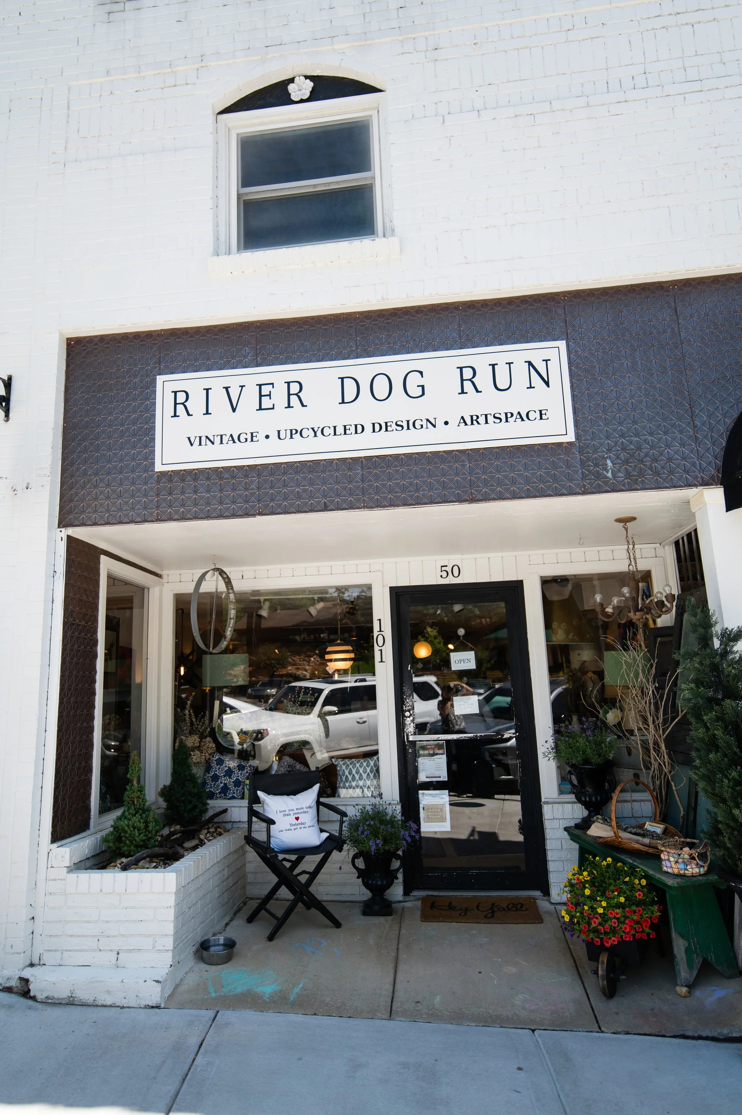 Storefront with sign reading 'RIVER DOG RUN, Vintage, Upcycled Design, Artspace.' The entrance has potted plants, a small white table, and decorative items.