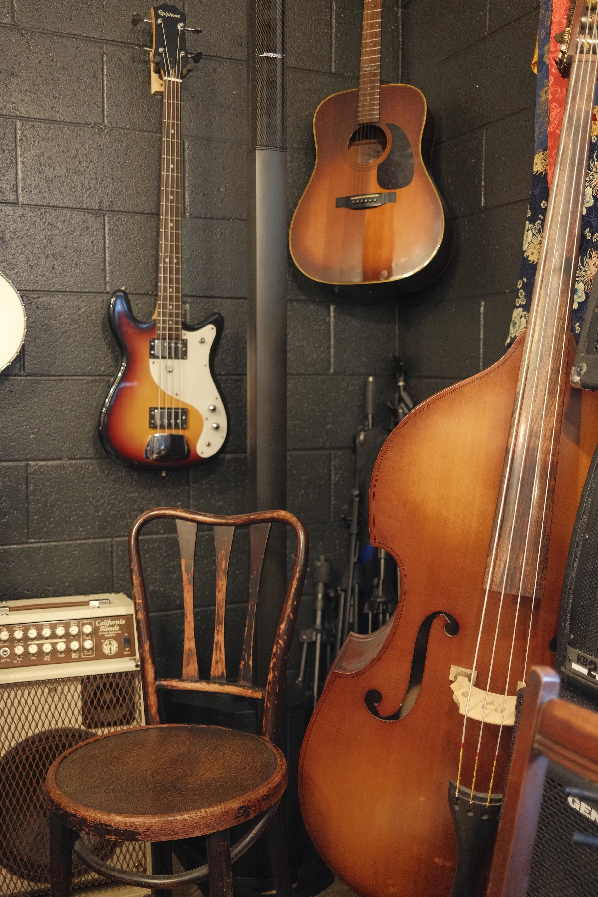 A room with musical instruments including a vintage bass guitar, a classical acoustic guitar, a double bass, and a small amplifier with a chair in front.