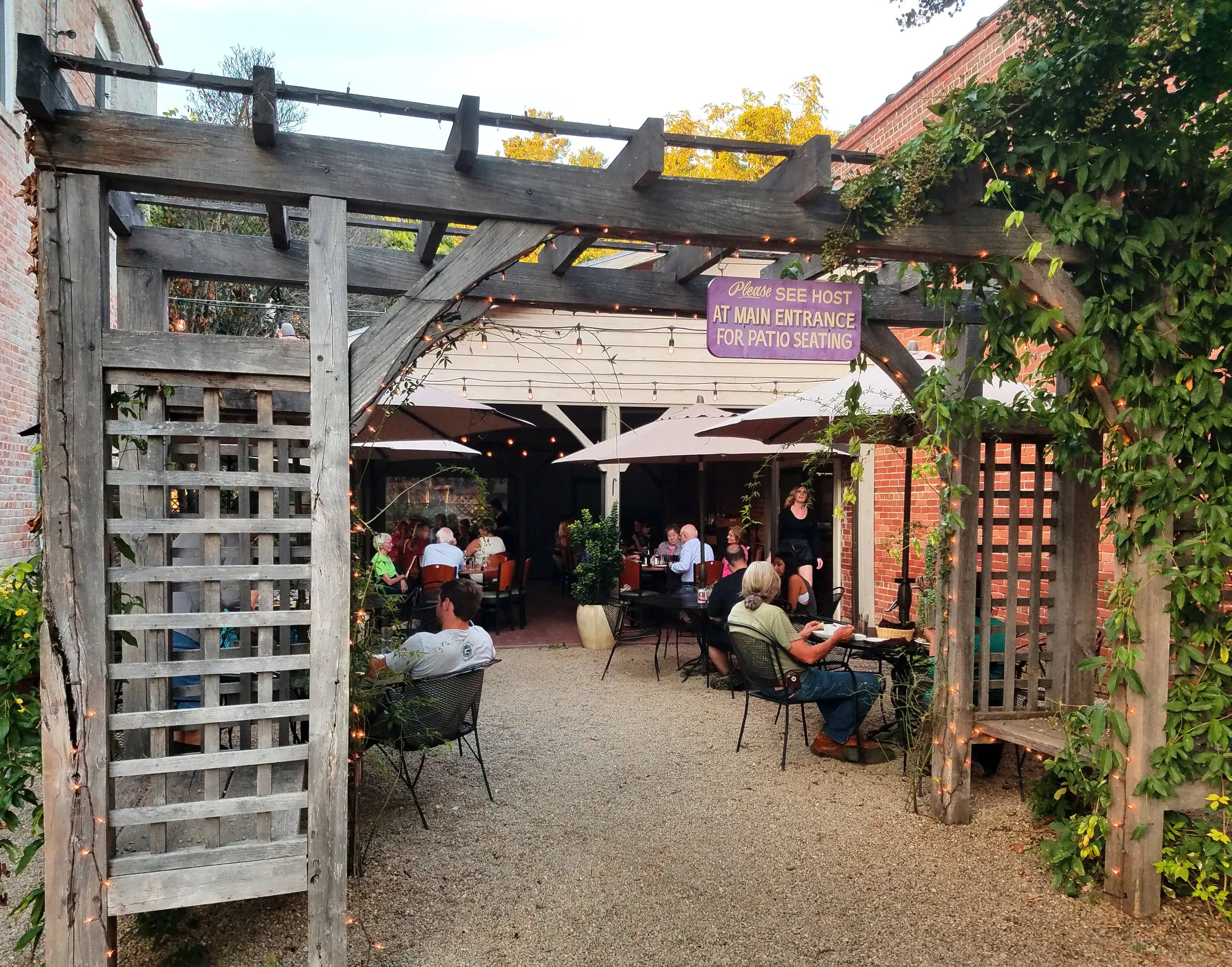An outdoor patio area with string lights, trellises, and tables with umbrellas, filled with people dining.