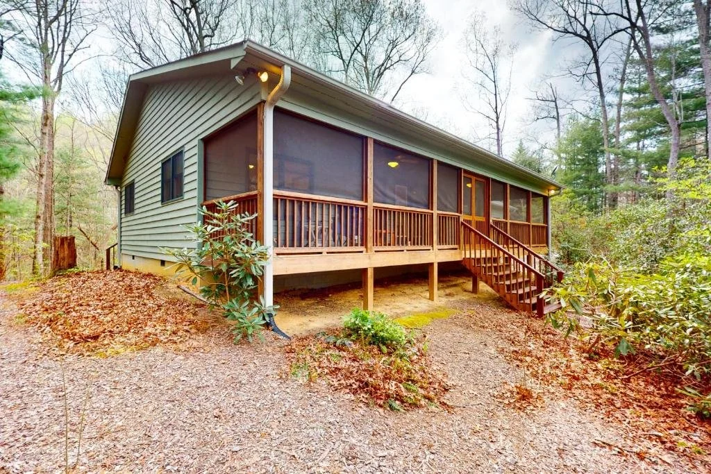 A house with a screened porch, situated in a wooded area with bare trees, is elevated on supports with stairs leading to the porch. The house has beige siding with dark trim.