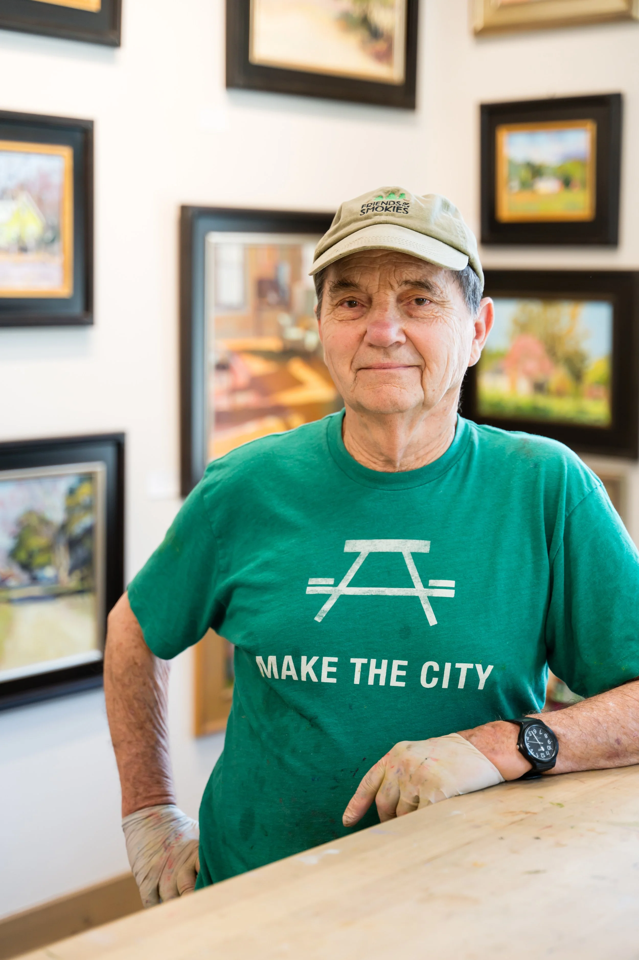 An elderly man wearing a beige cap and green t-shirt with a white graphic and text that reads 'MAKE THE CITY'. He is in an art gallery, standing at a table, smiling, with paintings on the wall behind him. His right arm is in a sling, and he wears a b